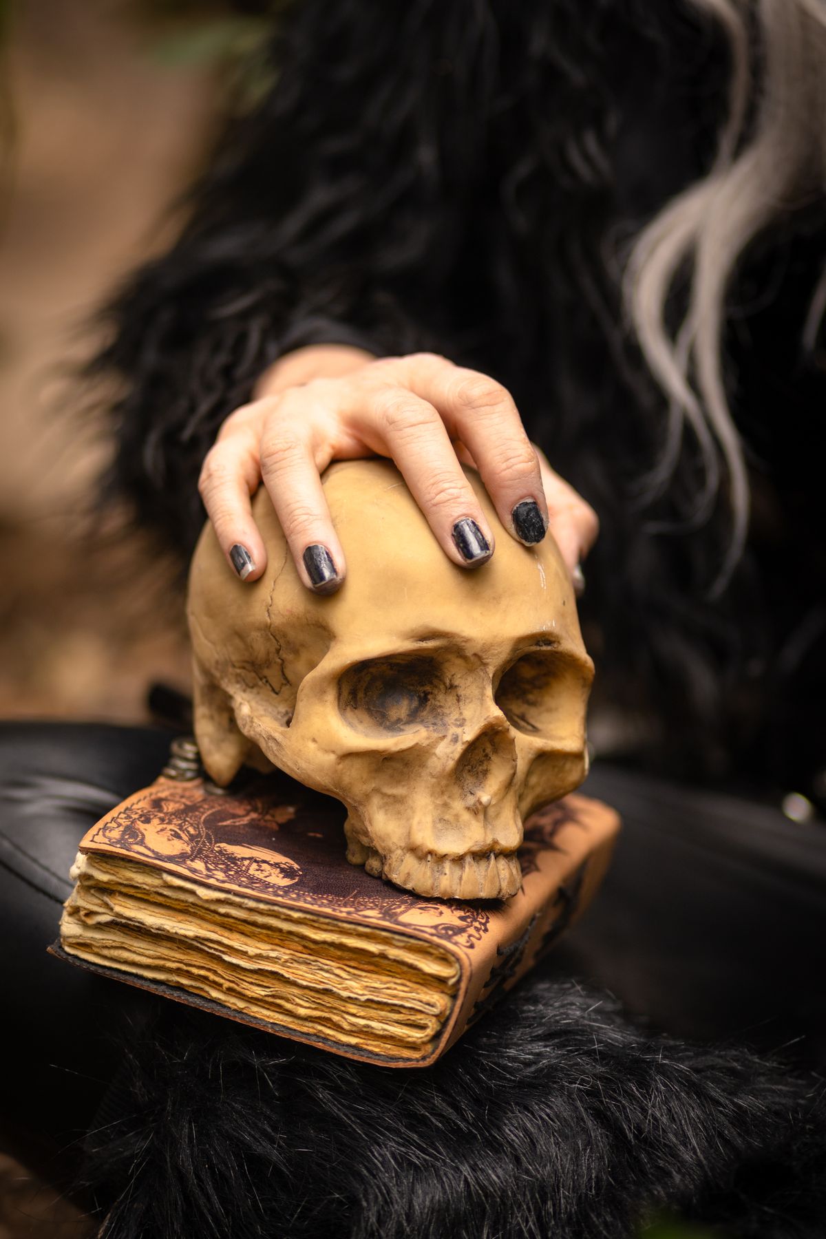Close-up of a hand with black nails holding a skull on an old book, evoking mystery.