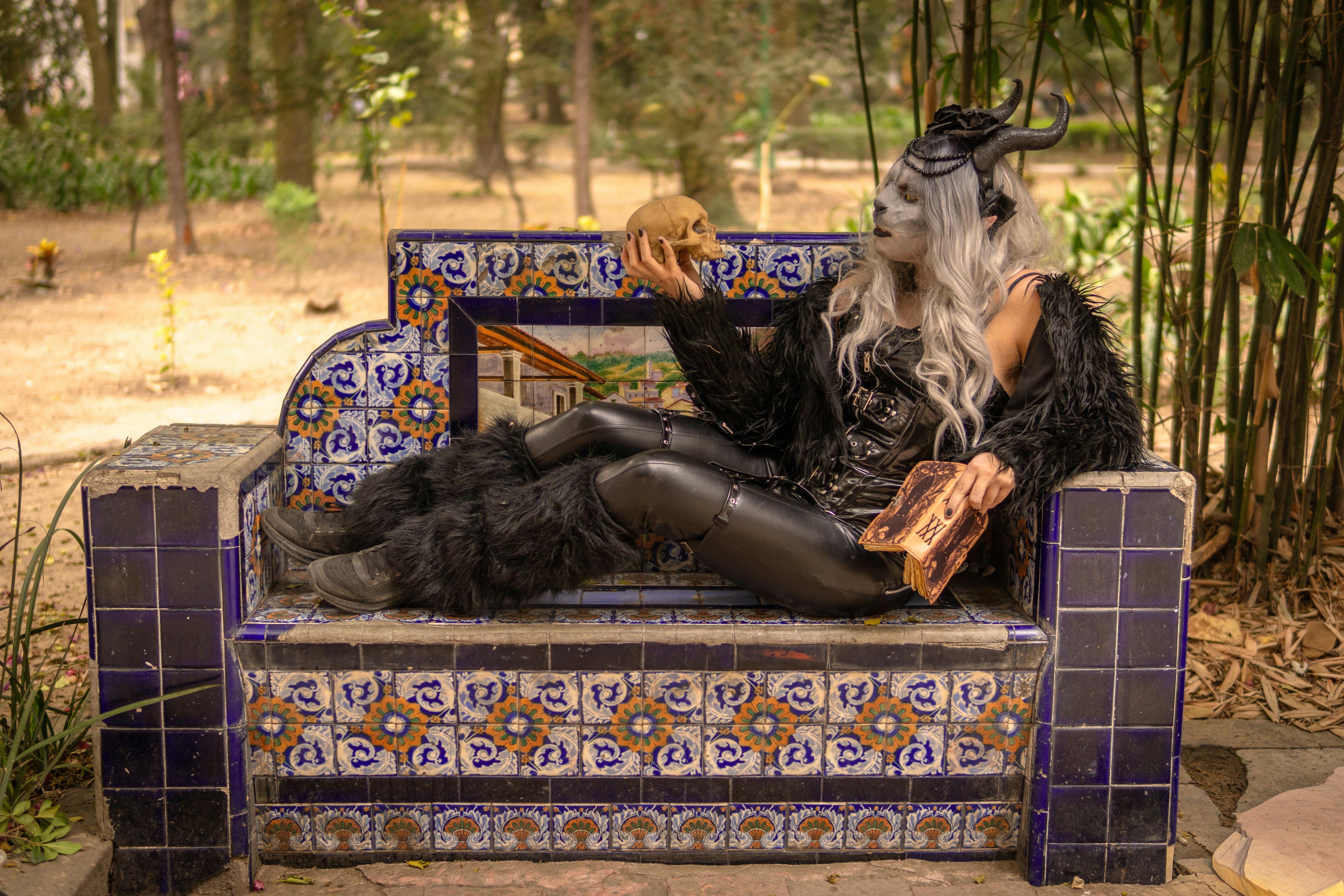 grátis Pessoa vestida com traje gótico, com chifres e caveira, sentada em um banco decorativo ao ar livre. Foto profissional