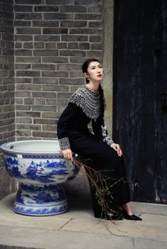 Elegant woman in ornate black dress beside traditional Chinese ceramic pot, posing indoors.