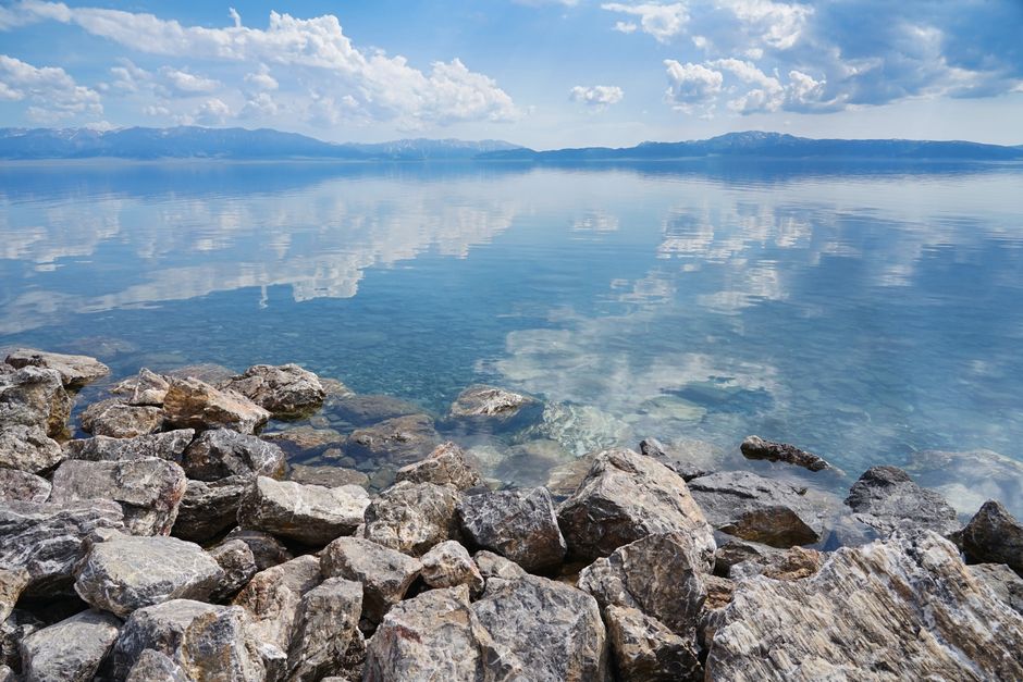 Tranquil lake with clear reflections of mountains and clouds surrounded by rocky shore under a blue sky.
