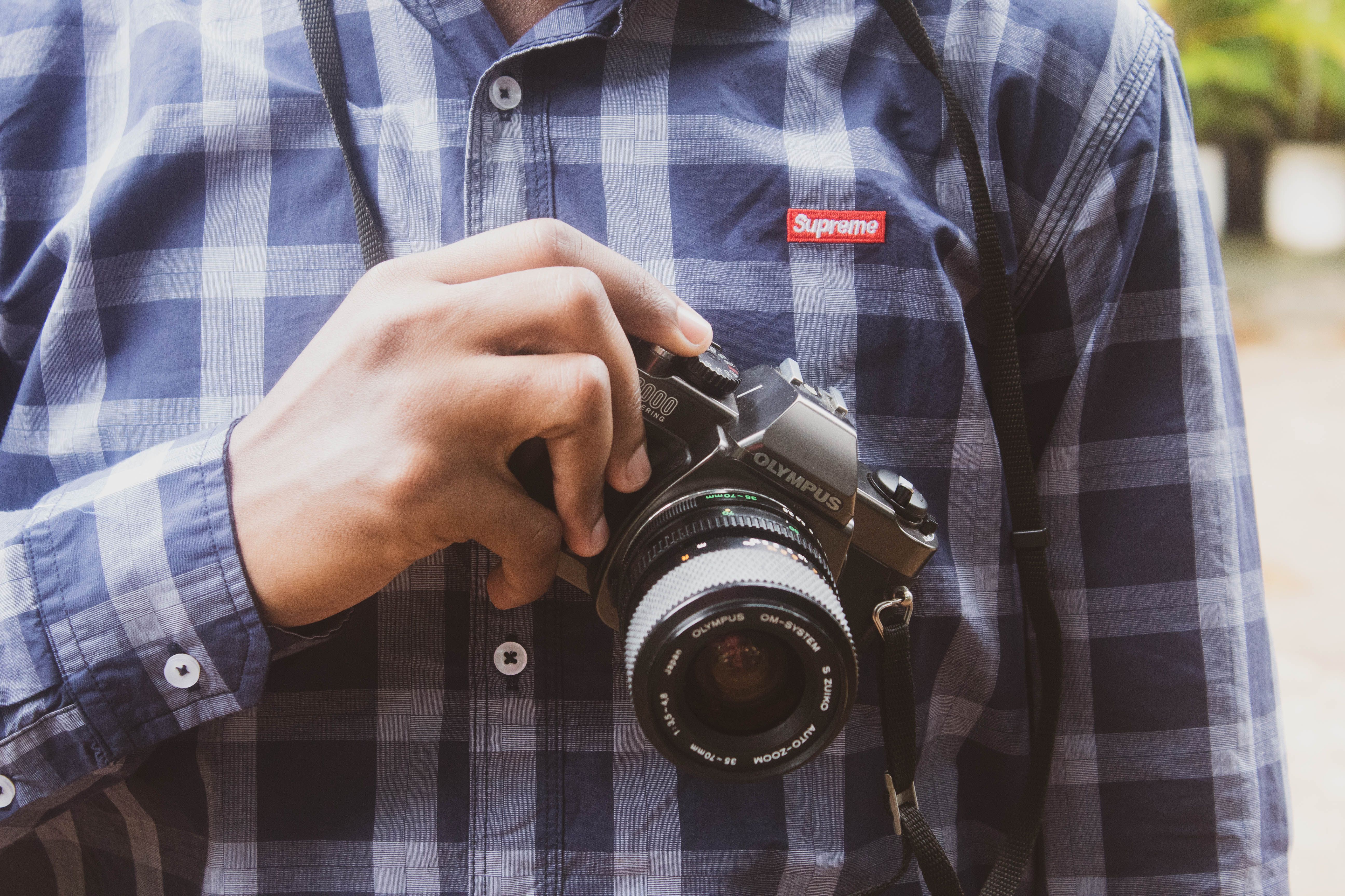 Close-up of a photographer holding an Olympus camera wearing a plaid shirt outdoors.