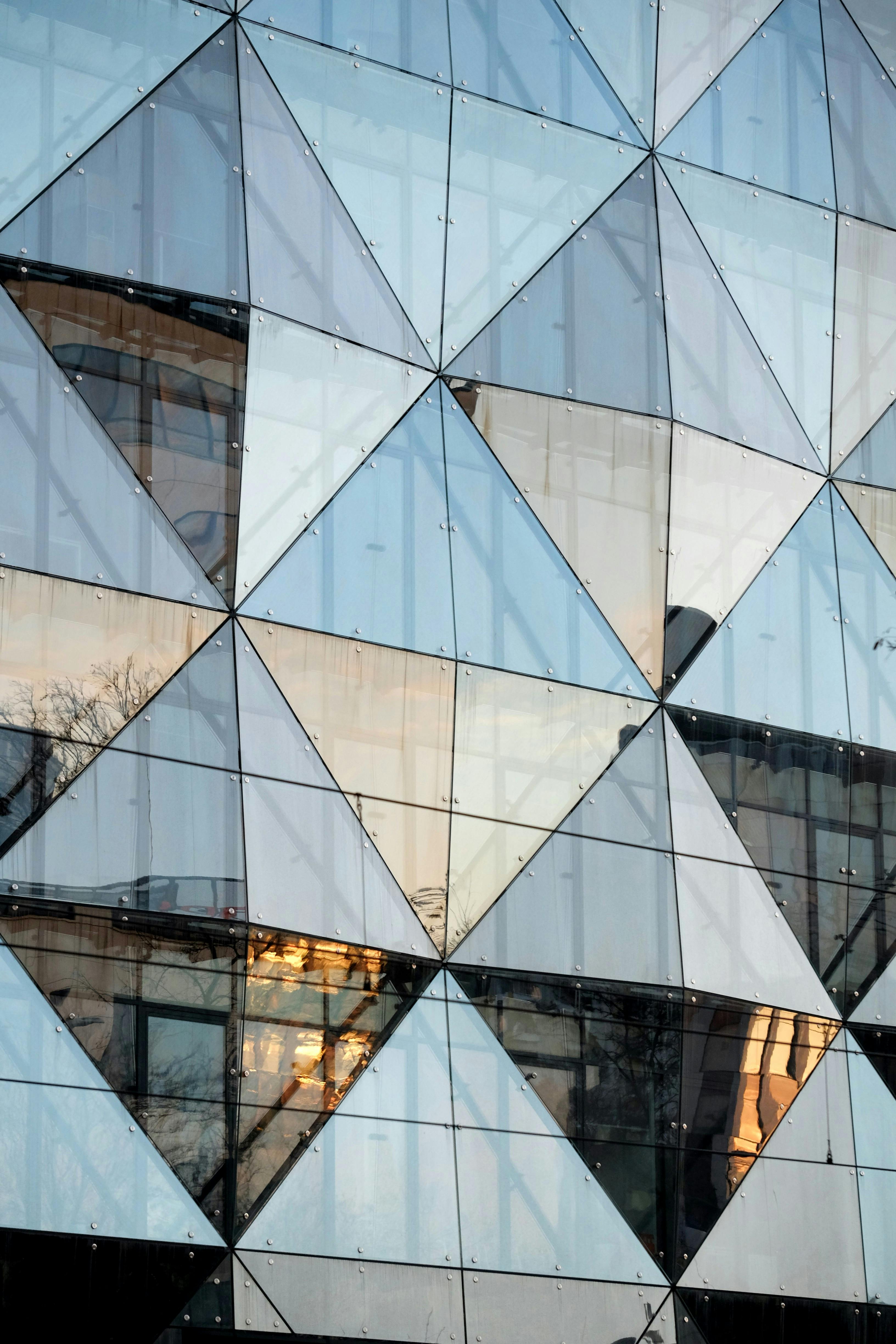 бесплатная Крупный план фасада современного стеклянного здания с геометрическими узорами, отражающими городской пейзаж. Стоковое фото