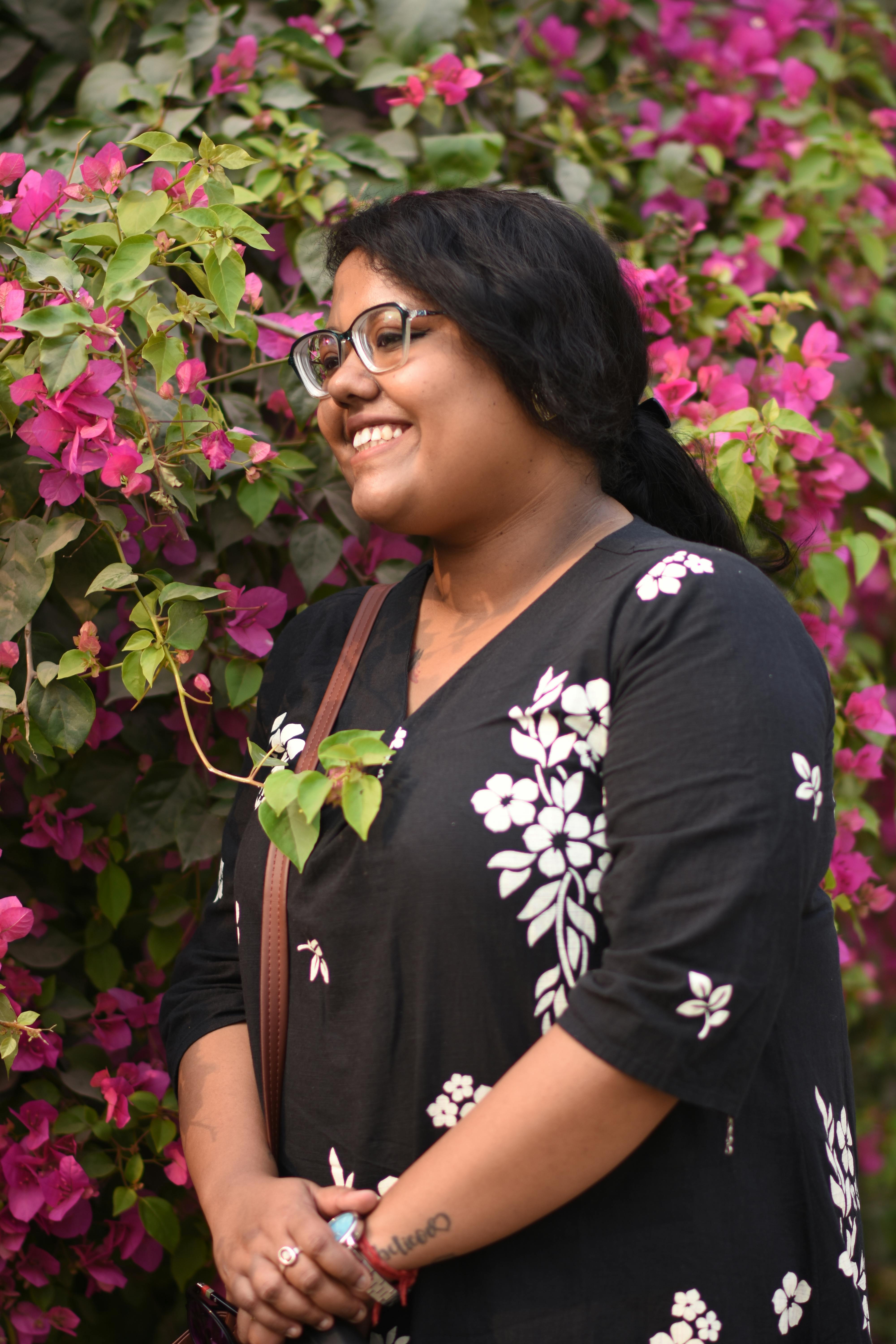 A woman smiles joyfully while standing beside blooming bougainvillea flowers, creating a vibrant and cheerful scene.