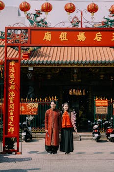 Couple in traditional attire stands outside a colorful Asian temple decorated with red lanterns.