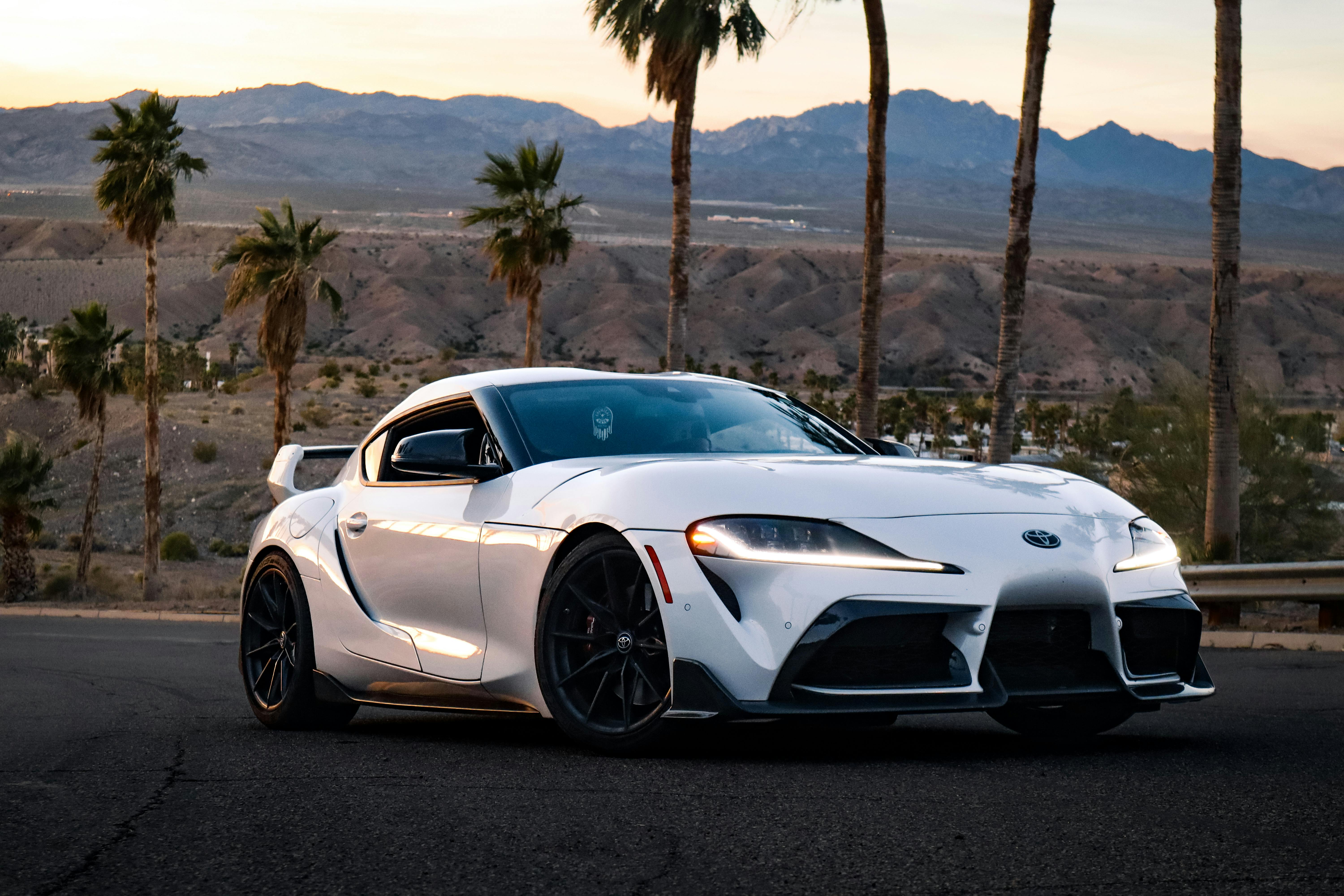 Gratis En hvit sportsbil parkert på en ørkenvei med utsikt over fjellene ved solnedgang. Arkivbilde