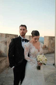 A stylish couple poses in elegant wedding attire outdoors at sunset.