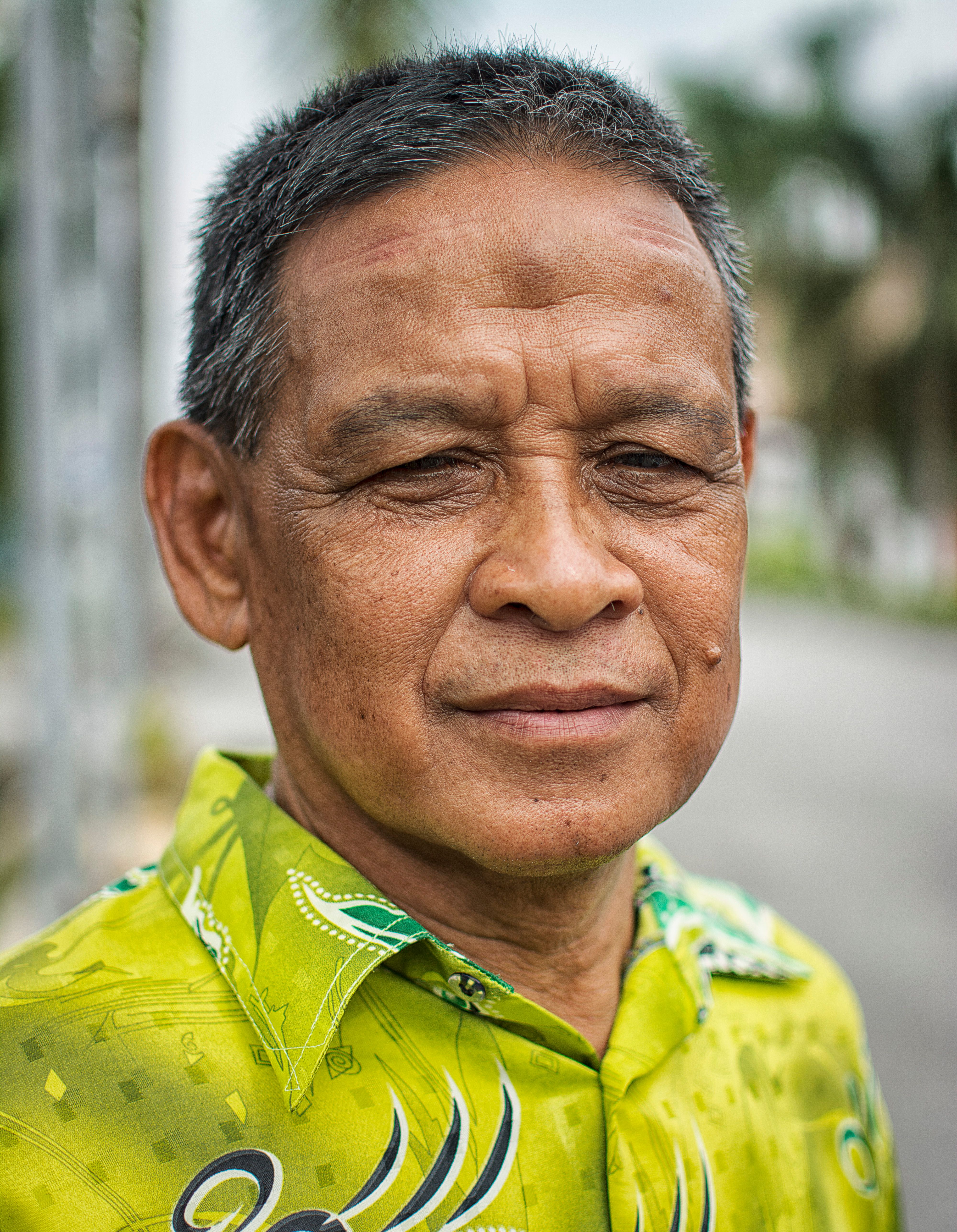 Portrait of an elderly man wearing a patterned green shirt, standing outdoors under natural light.