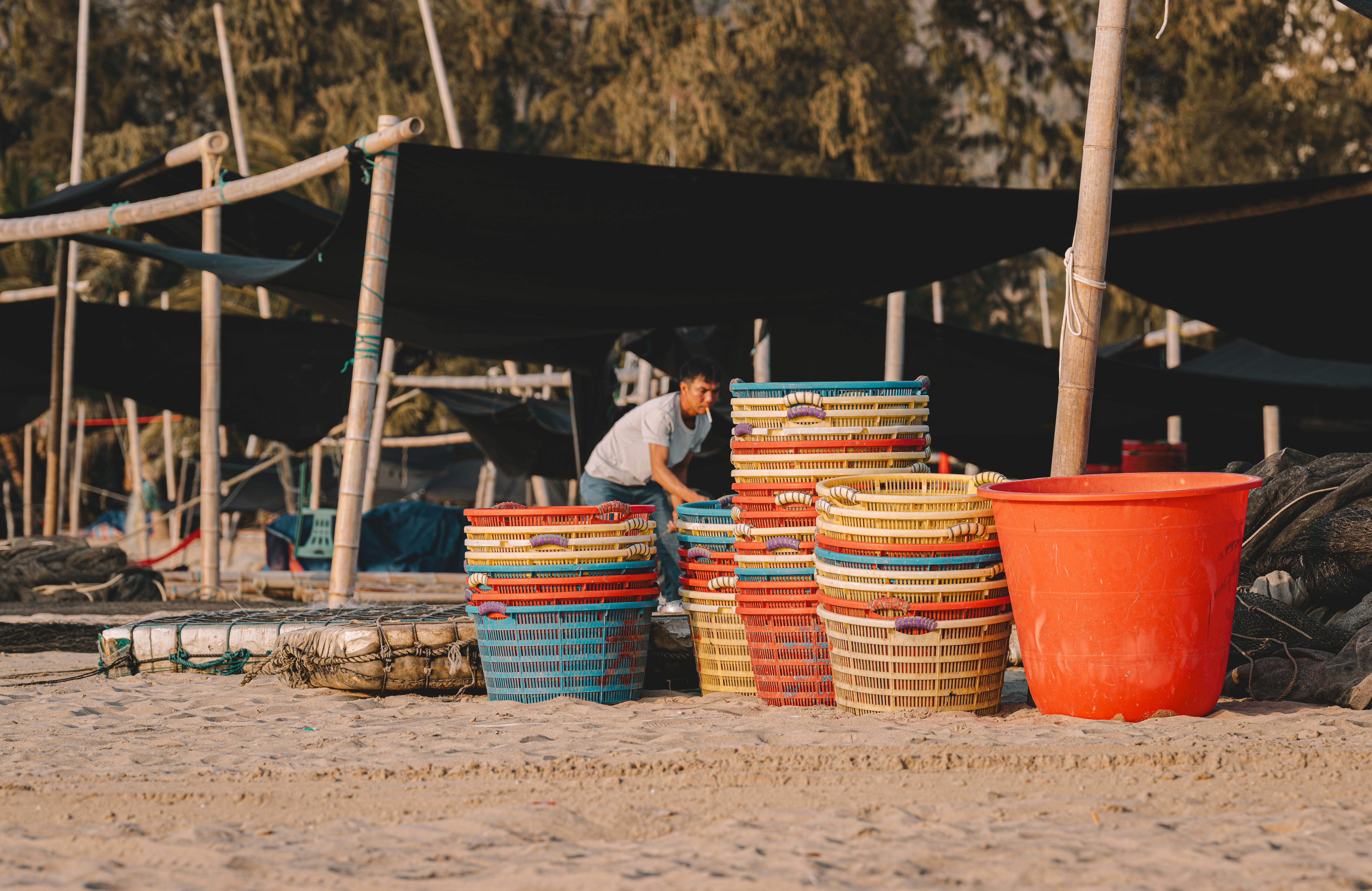 Gratis Seseorang menata keranjang-keranjang berwarna cerah di pantai yang diterangi matahari. Pemandangan pasar terbuka. Foto Stok