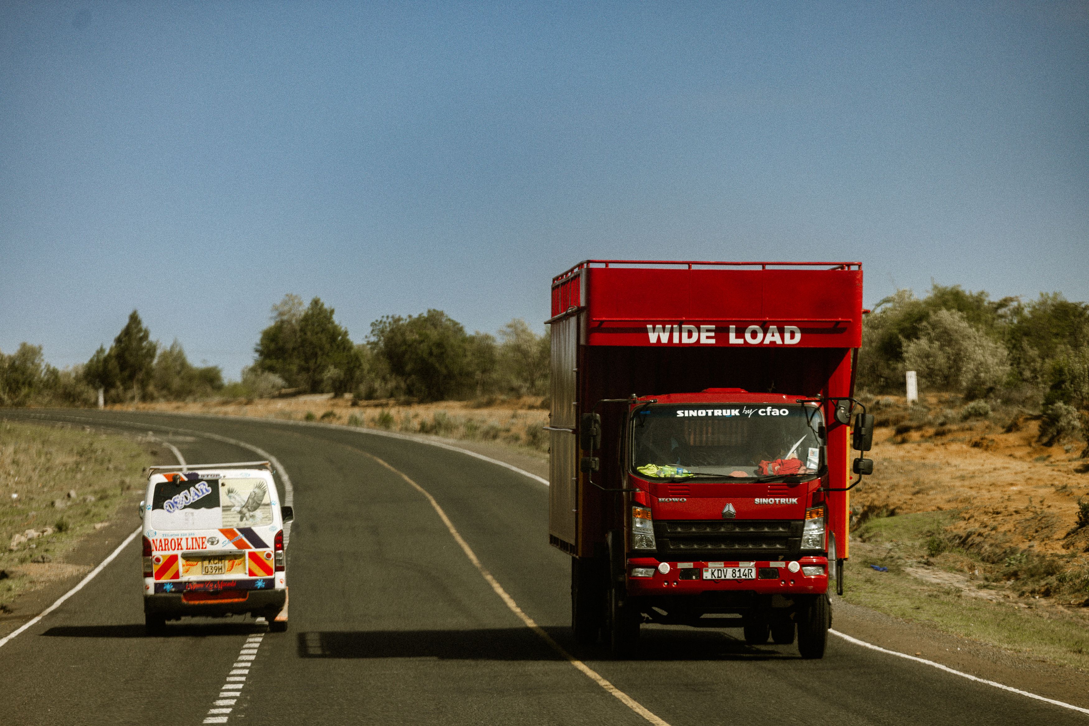 A red wide load truck and a van drive on an open highway through a desert landscape.
