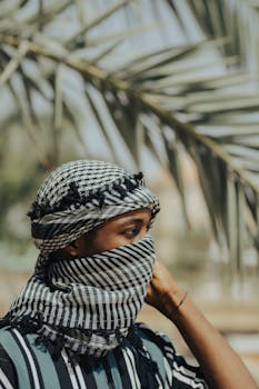 A person wearing traditional attire in Kano, Nigeria, standing outdoors with palm leaves in the background.
