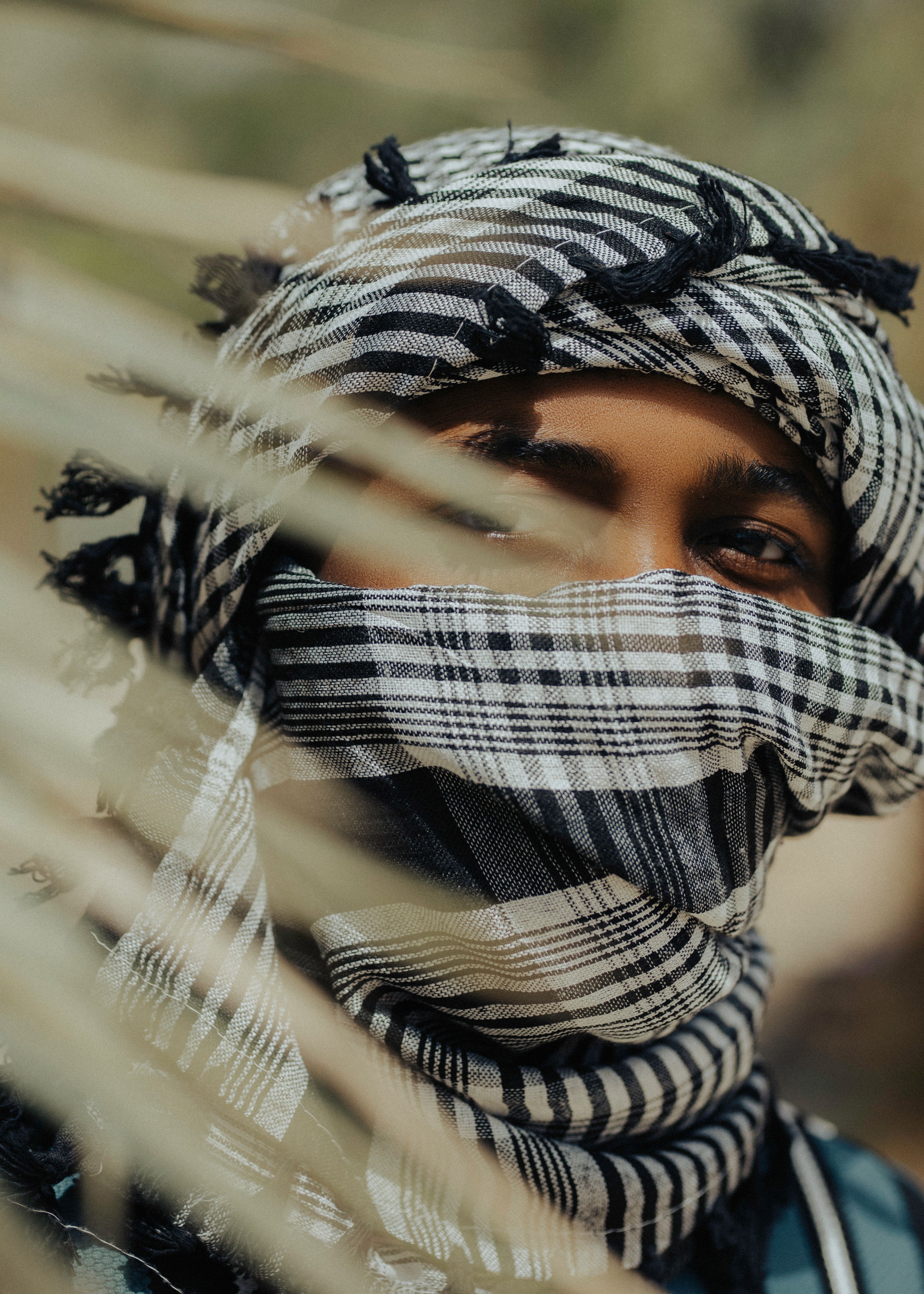 Close-up portrait of a man in traditional striped headscarf, partially hidden behind palm leaves outdoors in Kano, Nigeria.
