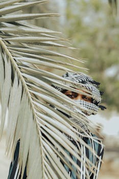 A man in traditional clothing peeks from behind a palm leaf outdoors in Kano, Nigeria.