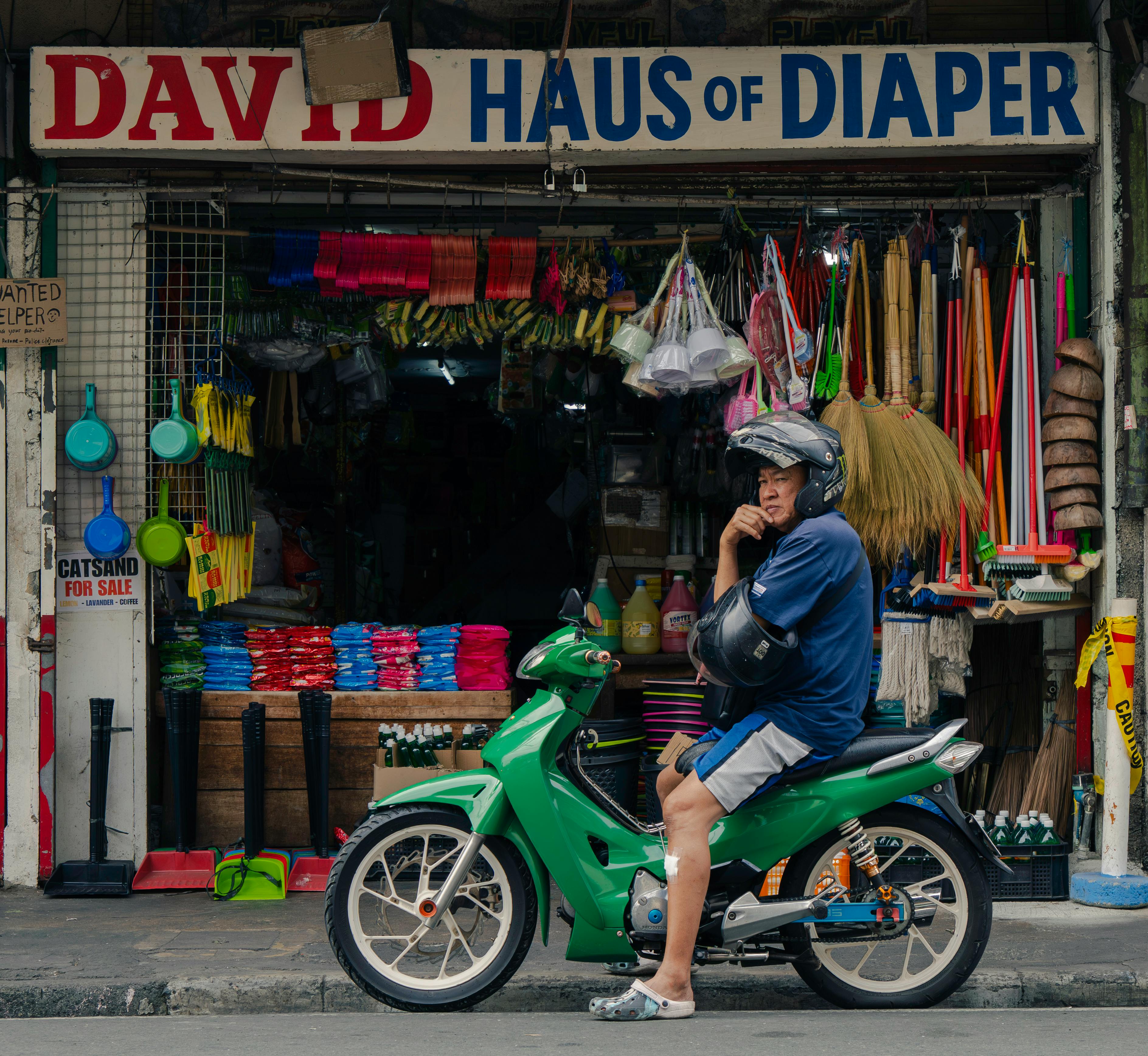 бесплатная Мужчина сидит на зелёном мотоцикле перед красочным магазином с товарами для дома. Стоковое фото