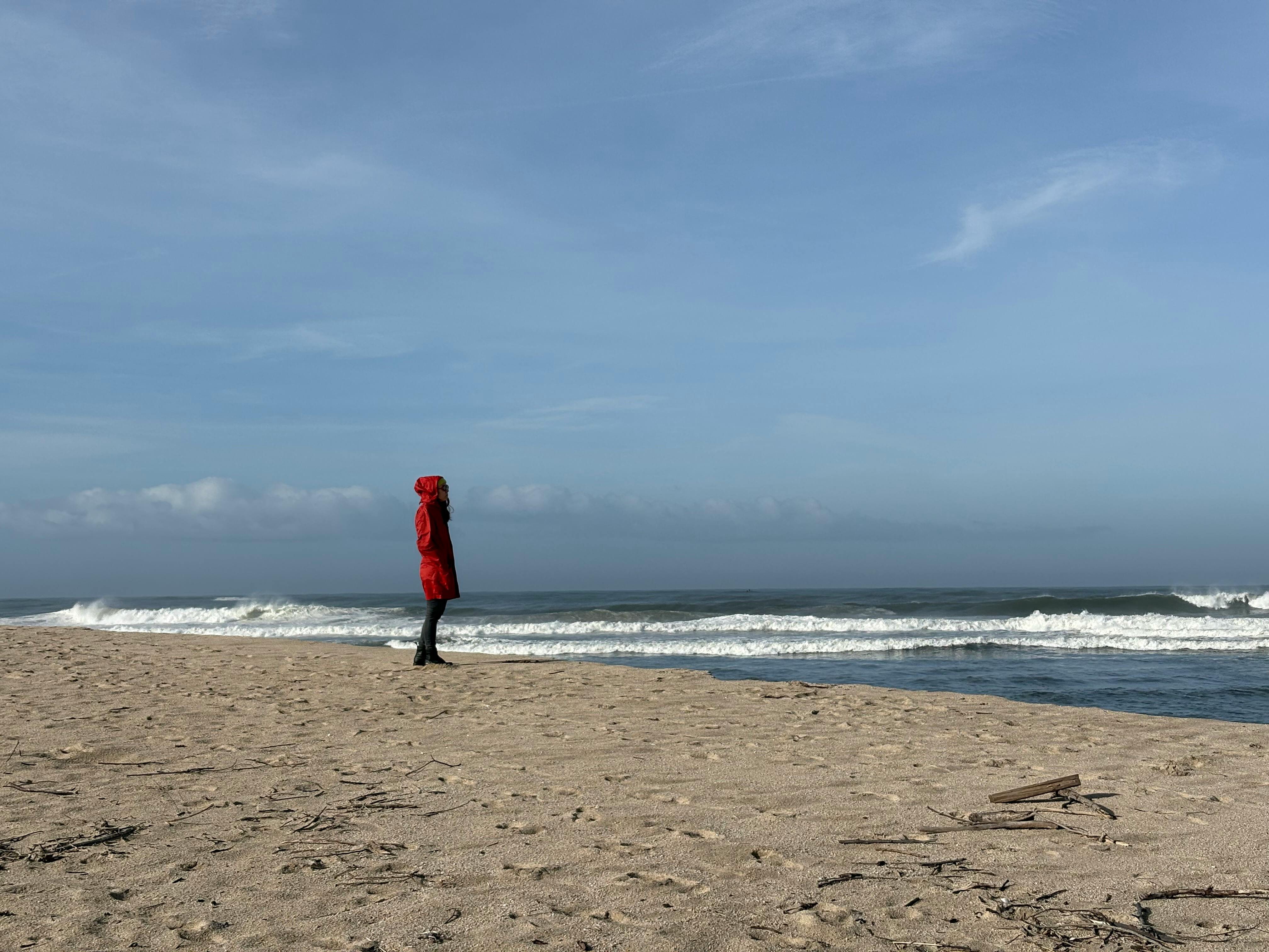 бесплатная На пляже Фао в Браге, Португалия, одинокая фигура в красном пальто стоит, глядя на океанские волны. Стоковое фото