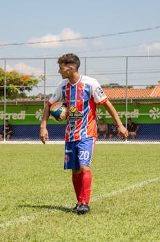 Soccer player in red and blue uniform holding a ball on a sunny day, ready for action.