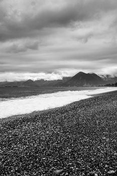 Black and white landscape of a pebble beach with waves and mountains under a cloudy sky.