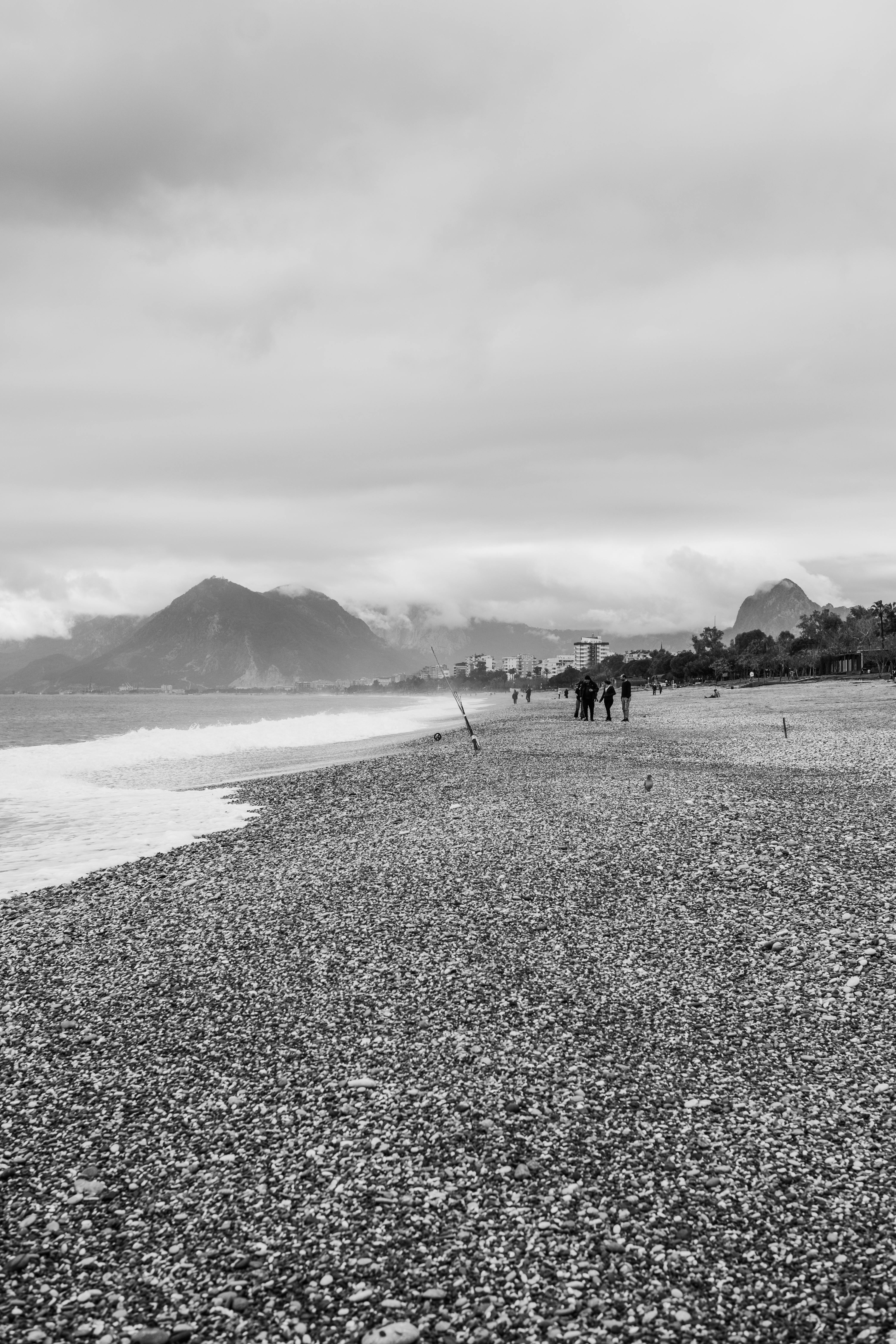 Gratis Pemandangan monokrom pantai berbatu dengan pegunungan di latar belakang dan orang-orang yang berjalan kaki. Foto Stok