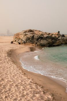 A serene view of a beach with a rocky outcrop, surrounded by gentle waves and distant city views.