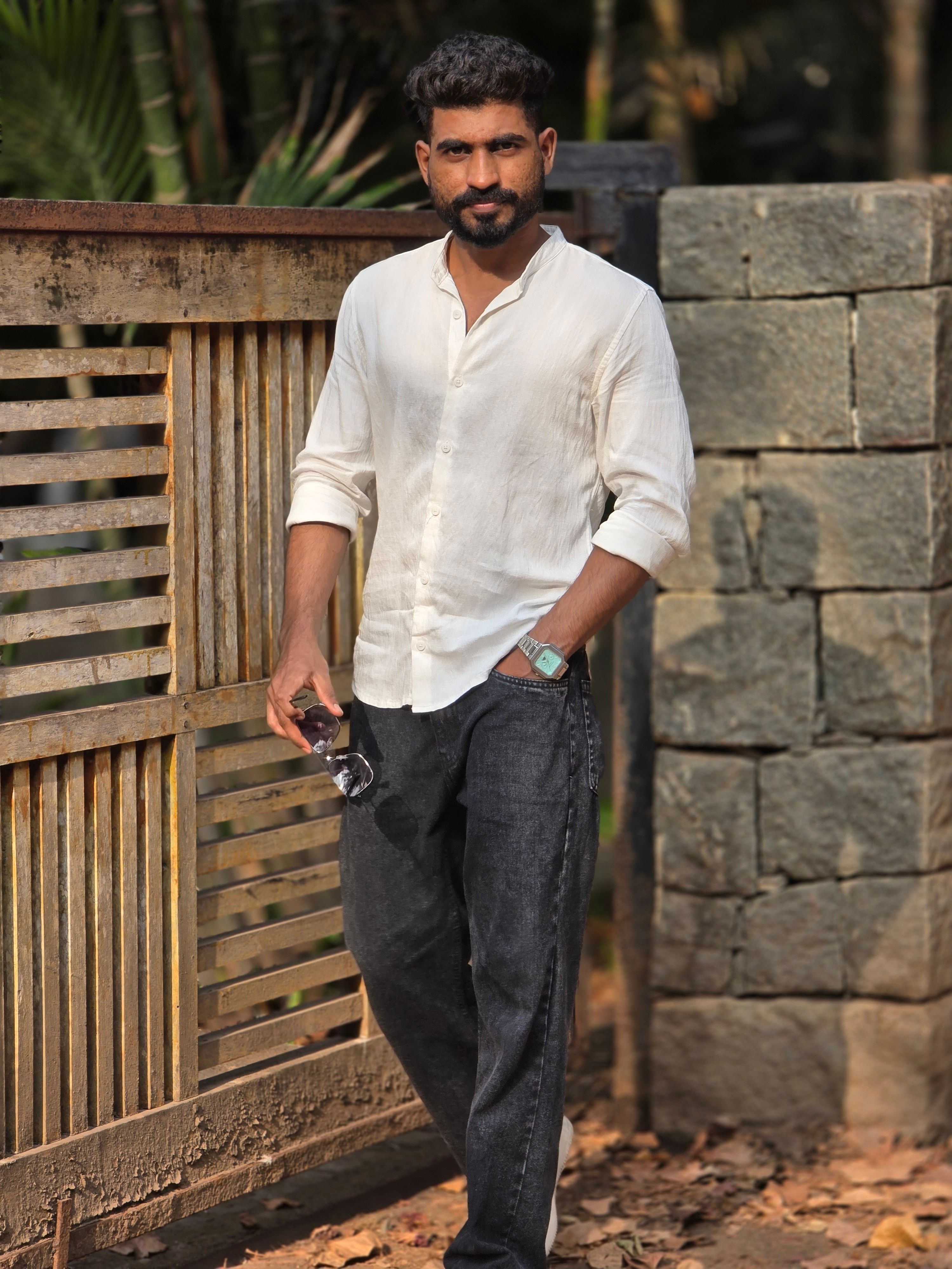 A confident man poses casually by a wooden and stone gate in natural outdoor light.