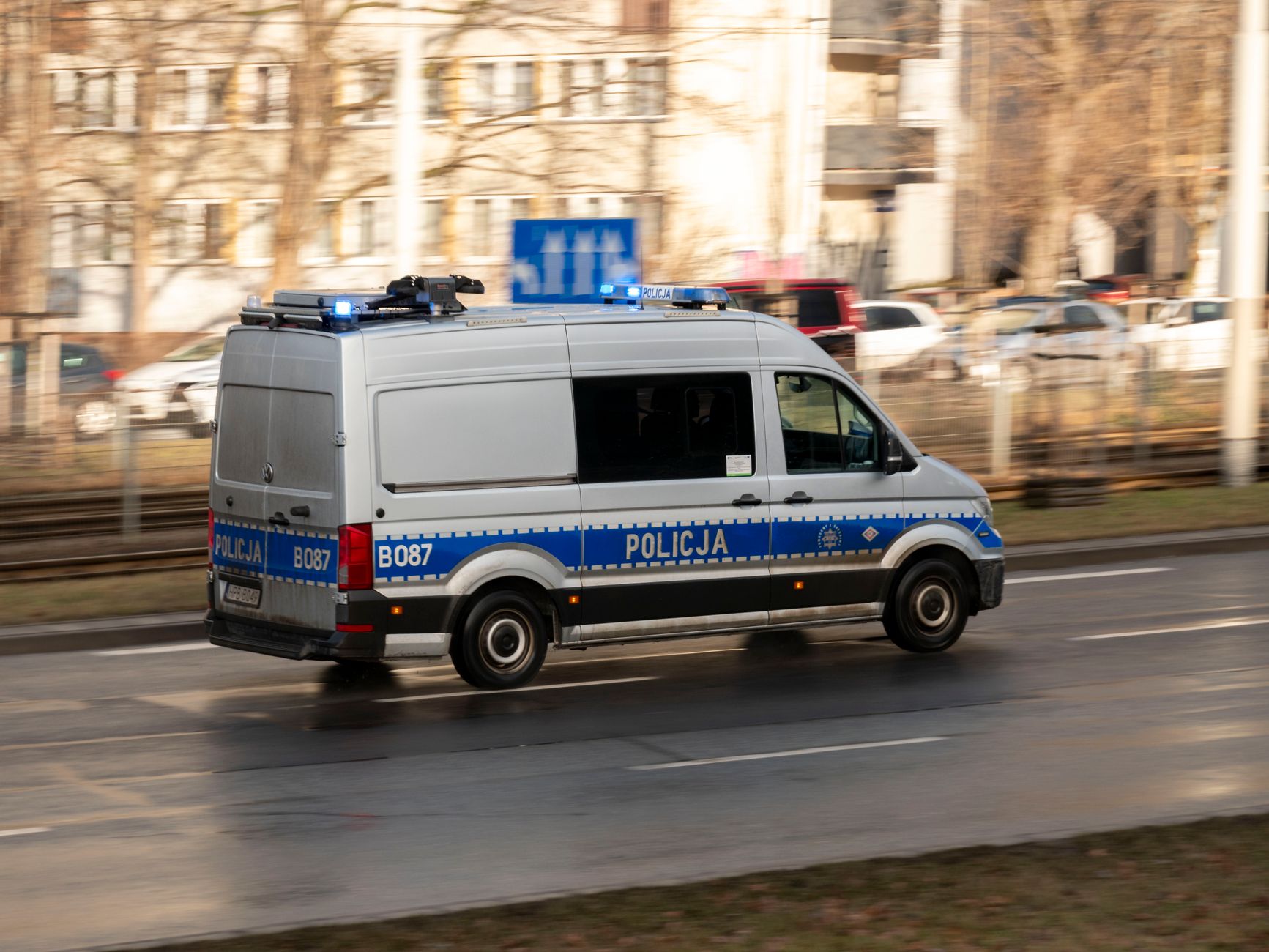 A Polish police van with blue lights on, driving swiftly on a city road.