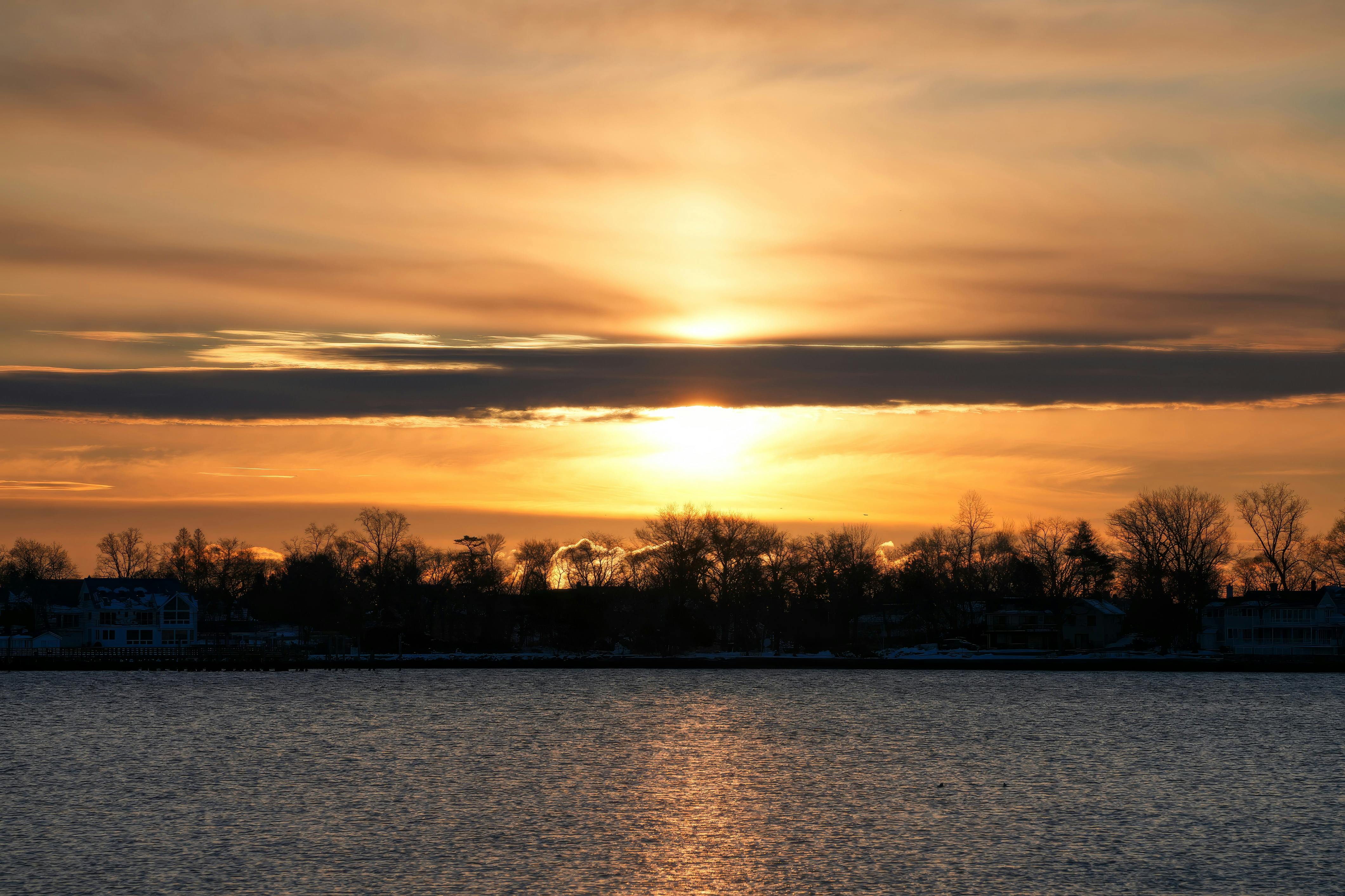 бесплатная Безмятежный закат над спокойным озером с силуэтами деревьев и зданий. Стоковое фото