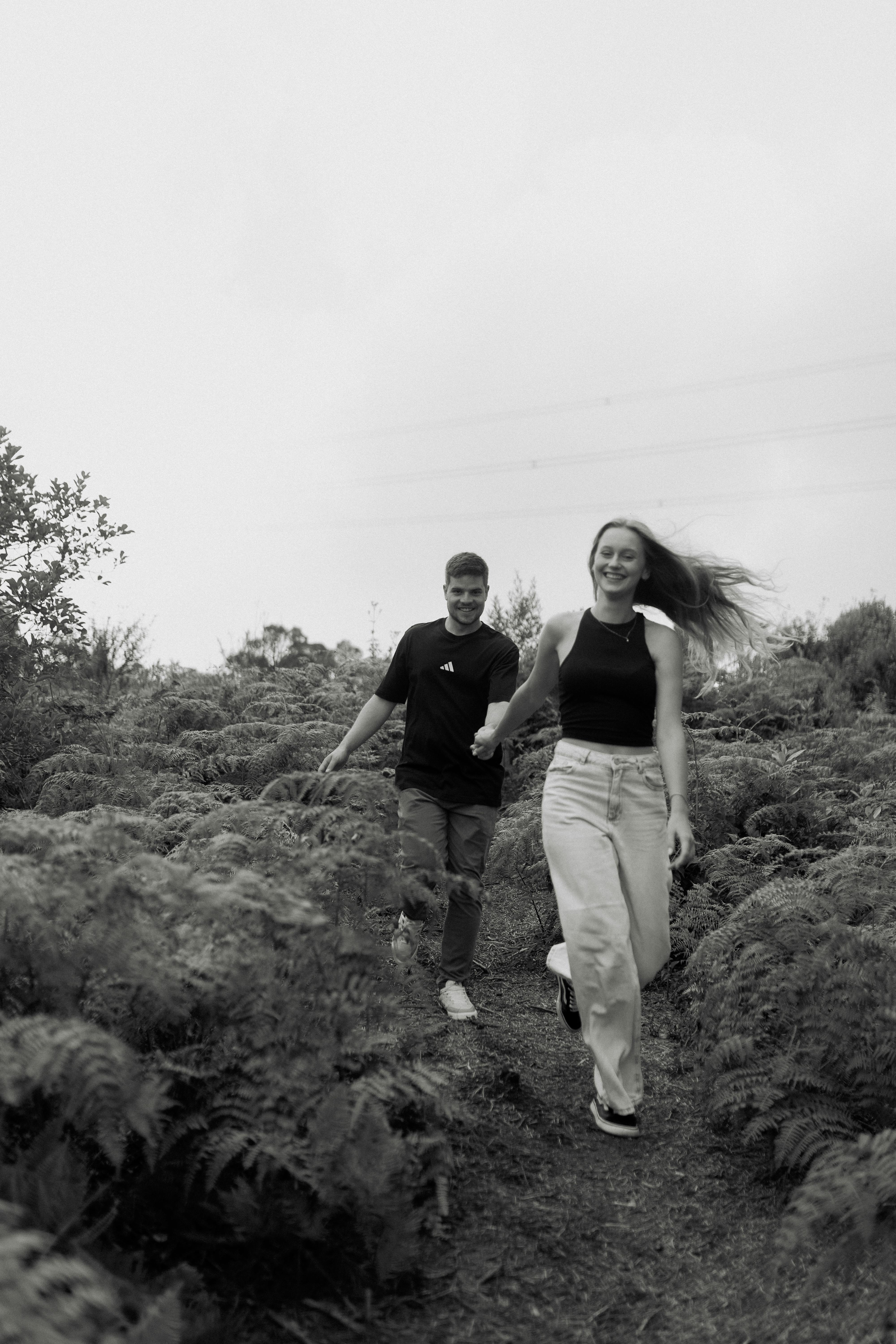 Gratis Sepasang kekasih yang bahagia berlari menyusuri jalur alam terbuka yang rimbun dalam foto hitam putih. Foto Stok