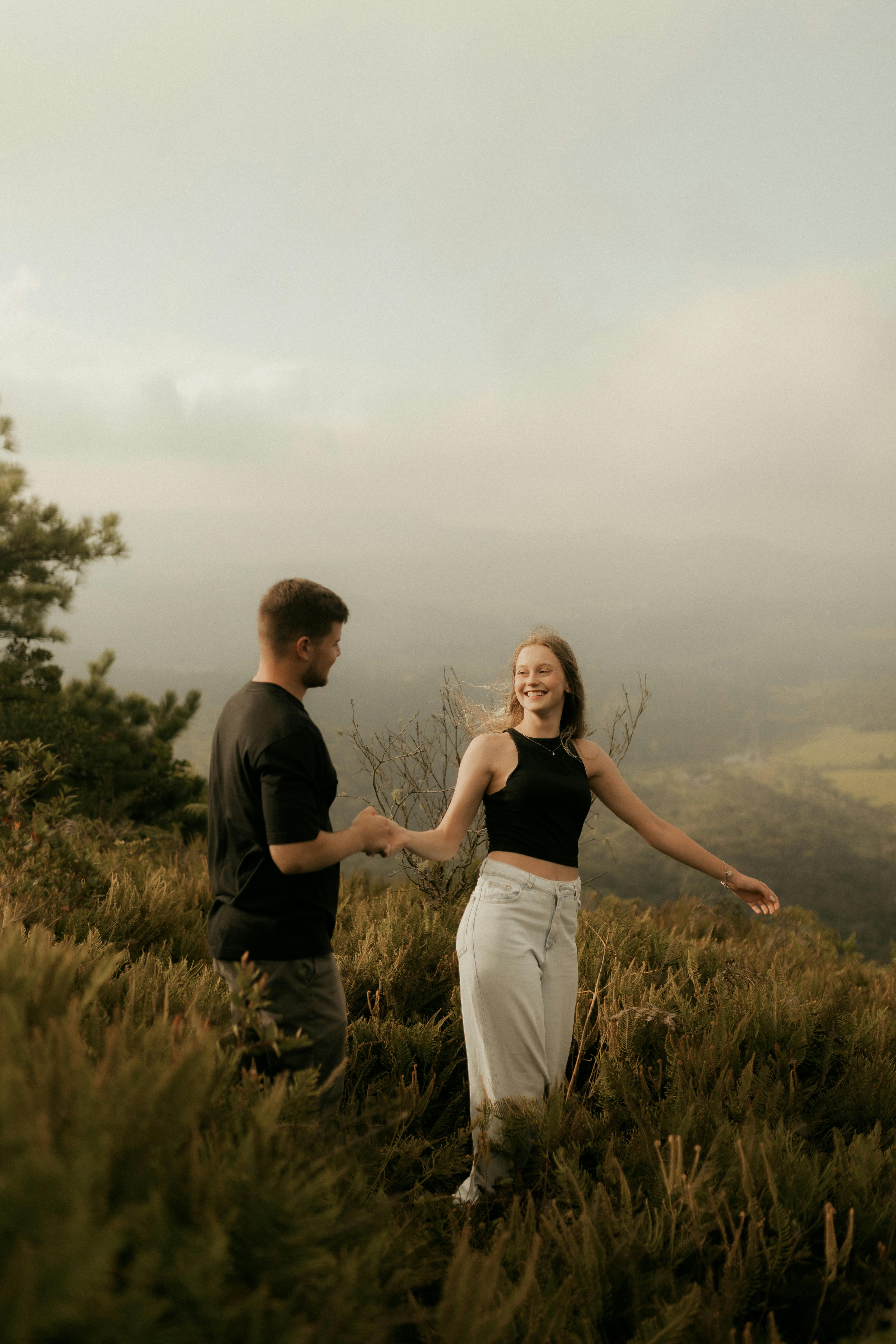Gratis Sepasang kekasih bergandengan tangan, tersenyum, di tengah lanskap hijau yang rimbun, menikmati pendakian yang indah. Foto Stok