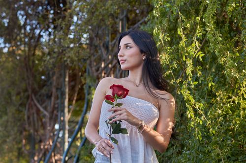 Free A woman stands outdoors holding a red rose, surrounded by lush greenery and basking in sunlight. Stock Photo