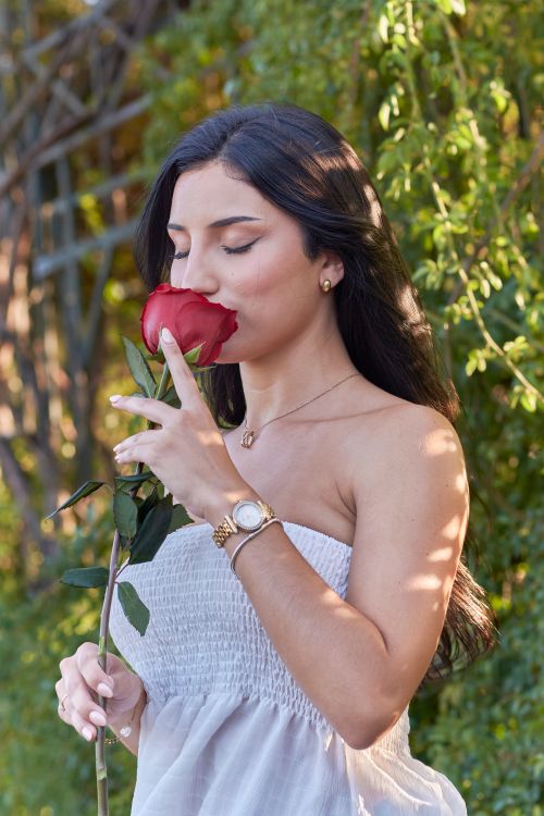 Free A young woman in a white dress enjoying the scent of a red rose in a bright garden setting. Stock Photo