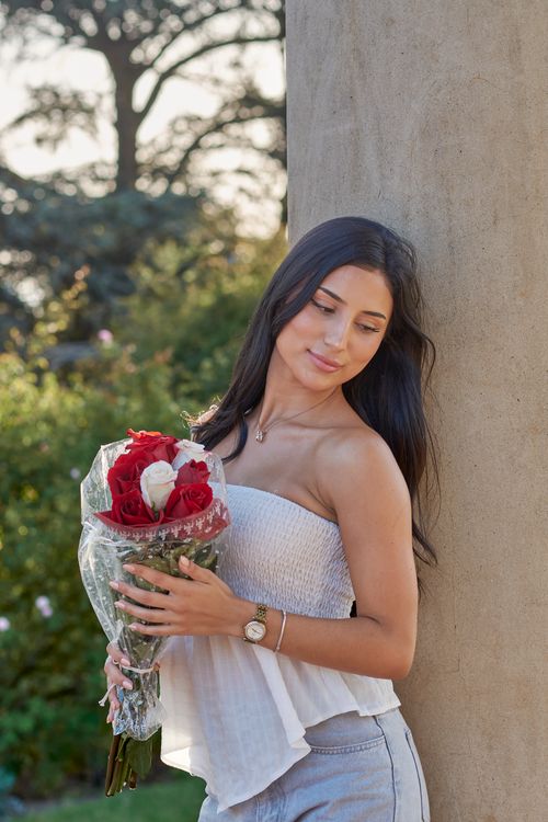 Free A woman leans against a column holding a bouquet of red roses, set in a lush garden in Montevideo. Stock Photo