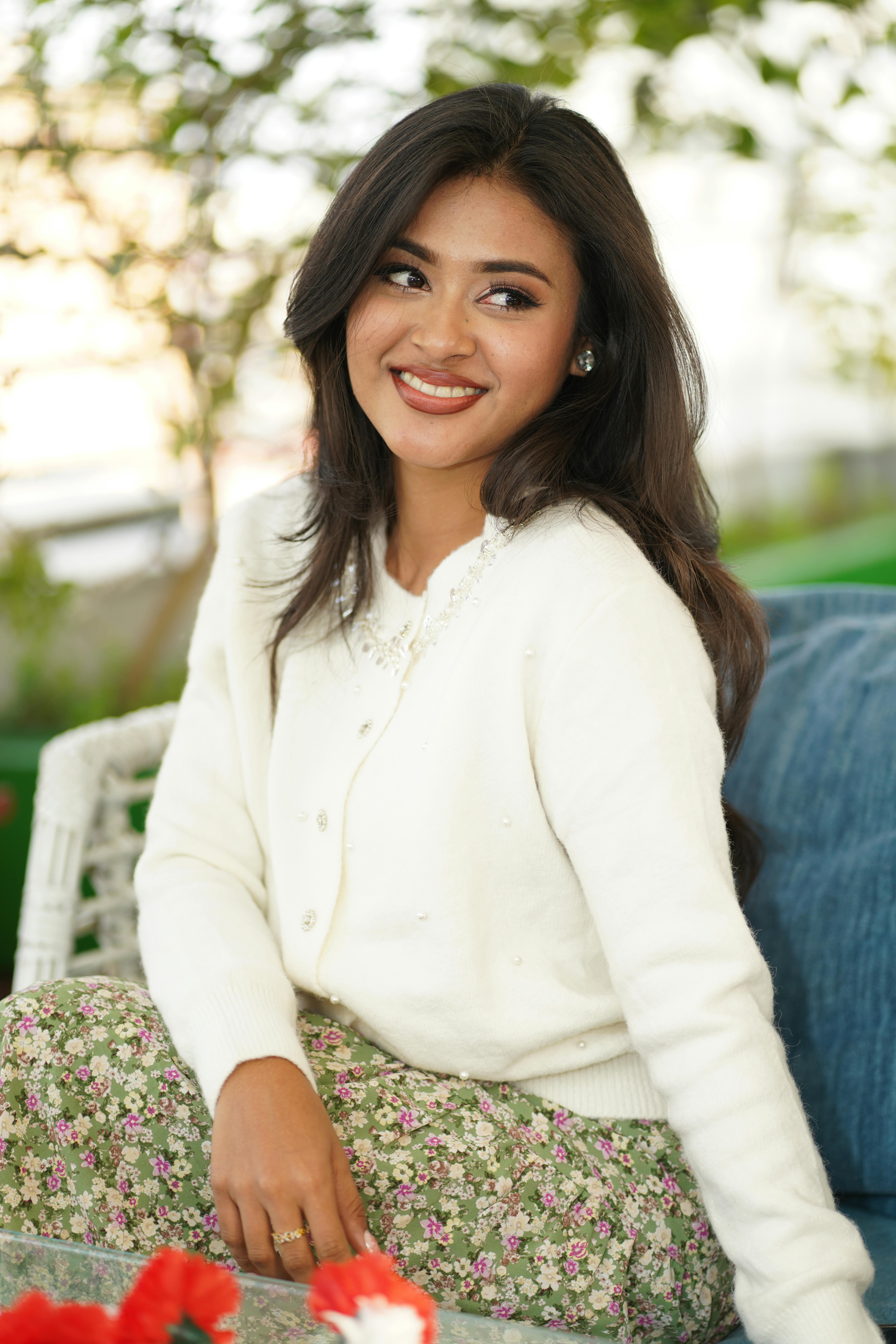 Gratis Portrett av en smilende kvinne som sitter utendørs i en hvit genser og et blomstrete skjørt, som fanger en avslappet og munter stemning. Arkivbilde