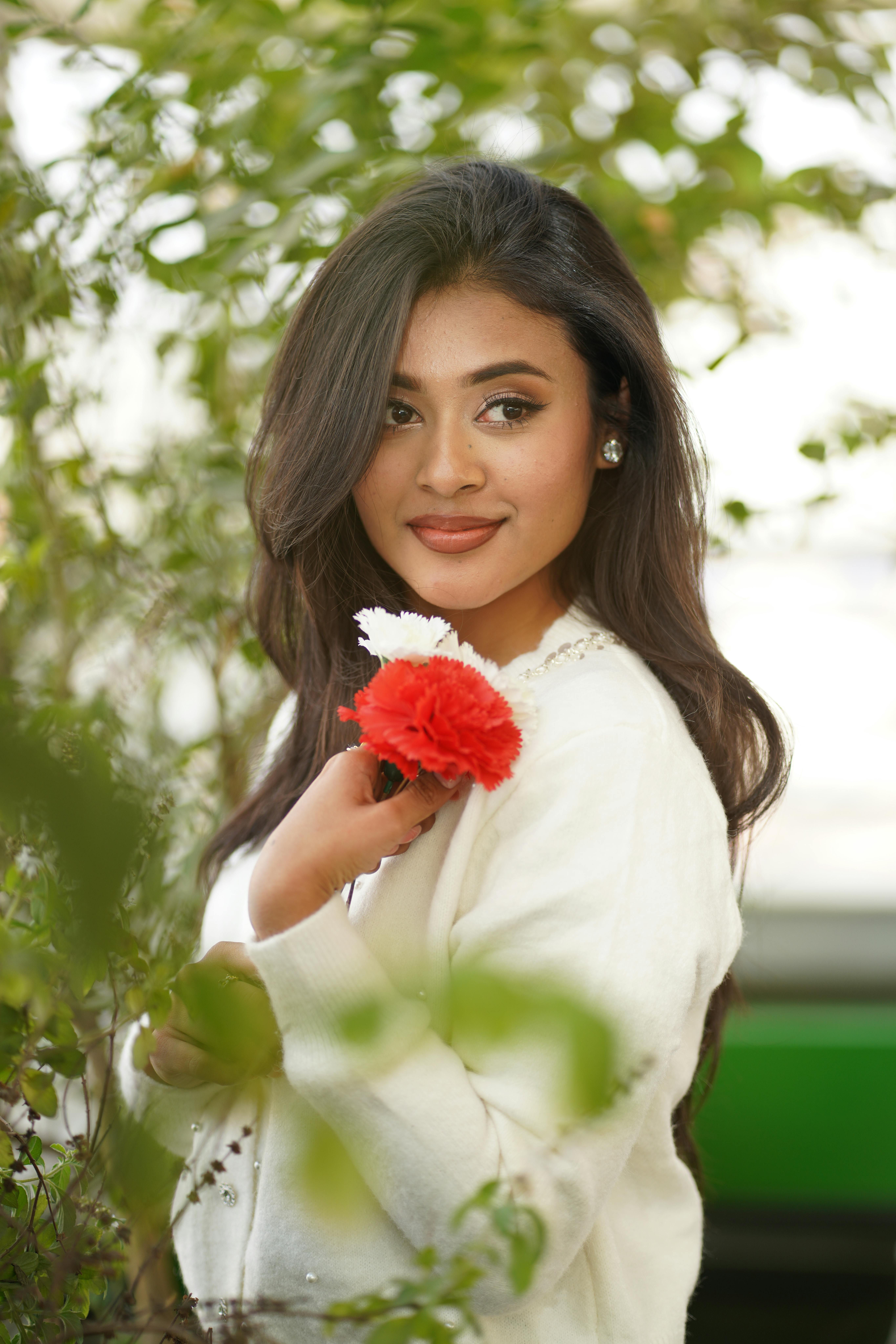 Gratis En kvinne i en hvit genser som holder røde og hvite blomster, omgitt av grøntområder. Arkivbilde