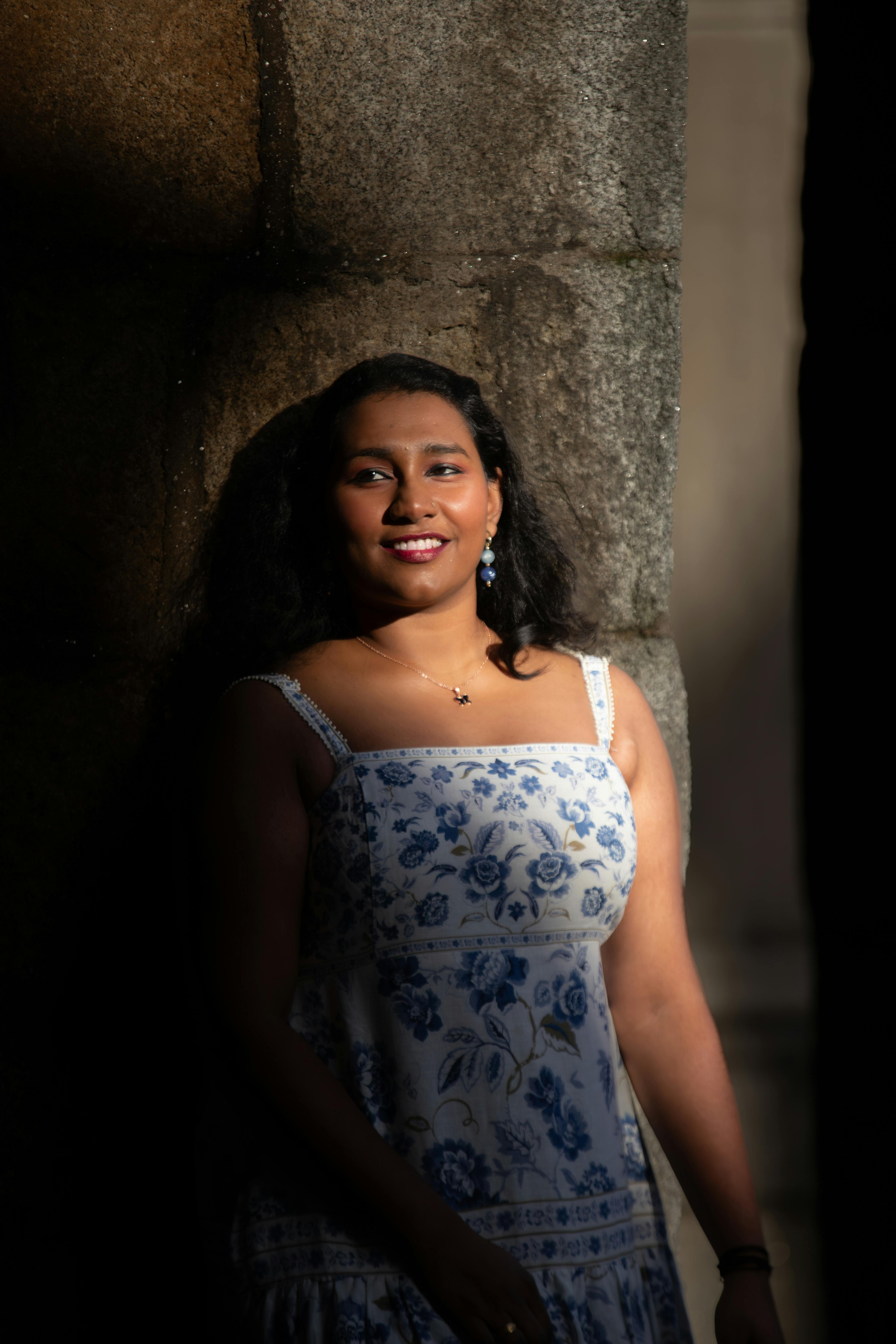 Gratis Seorang wanita tersenyum anggun mengenakan gaun bermotif bunga di bawah cahaya alami musim panas di dekat dinding batu. Foto Stok