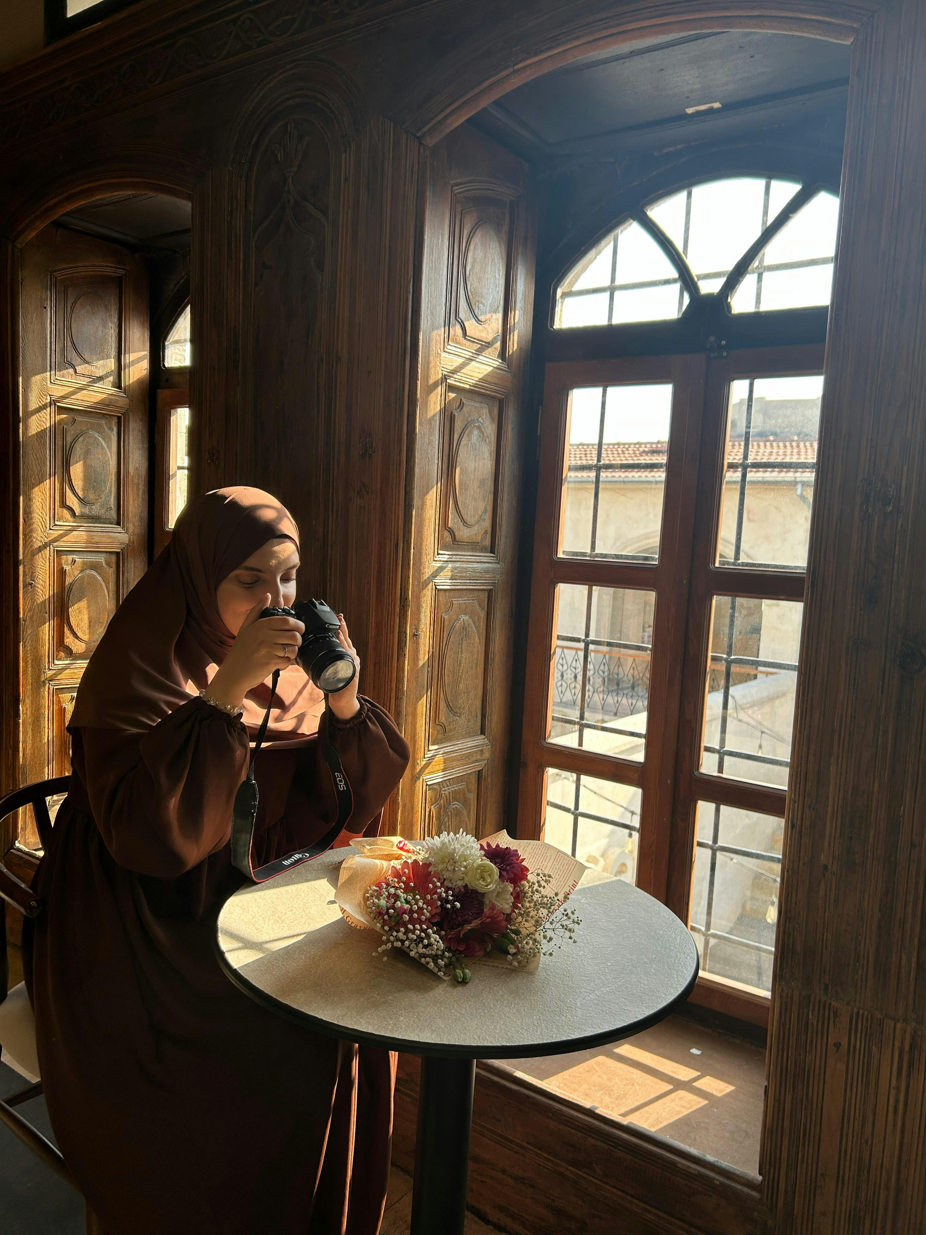 бесплатная Женщина в коричневой одежде пьет кофе у залитого солнцем окна, на столе стоит букет цветов. Стоковое фото