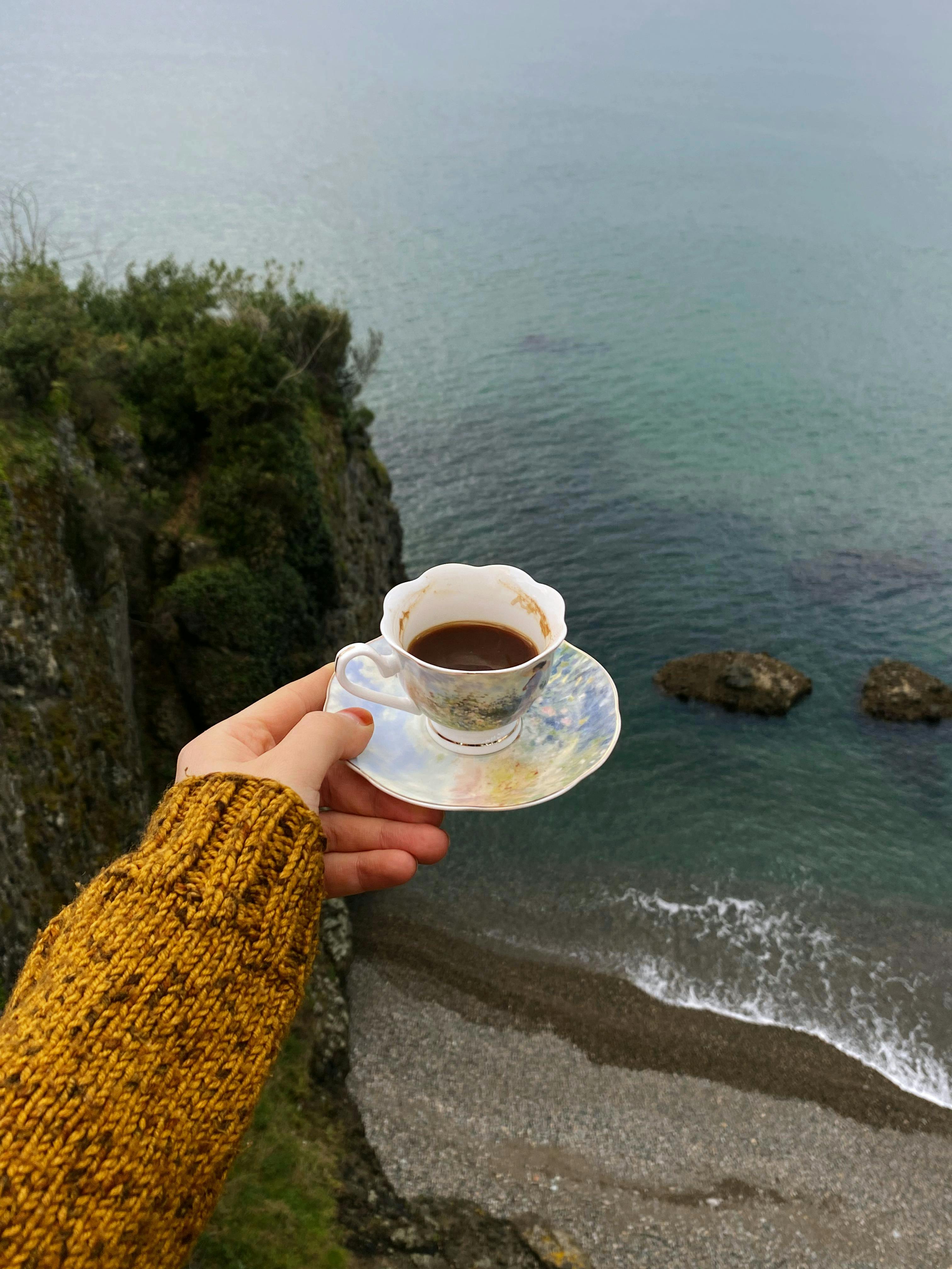 Gratis Nyt en fredelig utsikt over sjøen med en kopp kaffe holdt over klippen. Arkivbilde