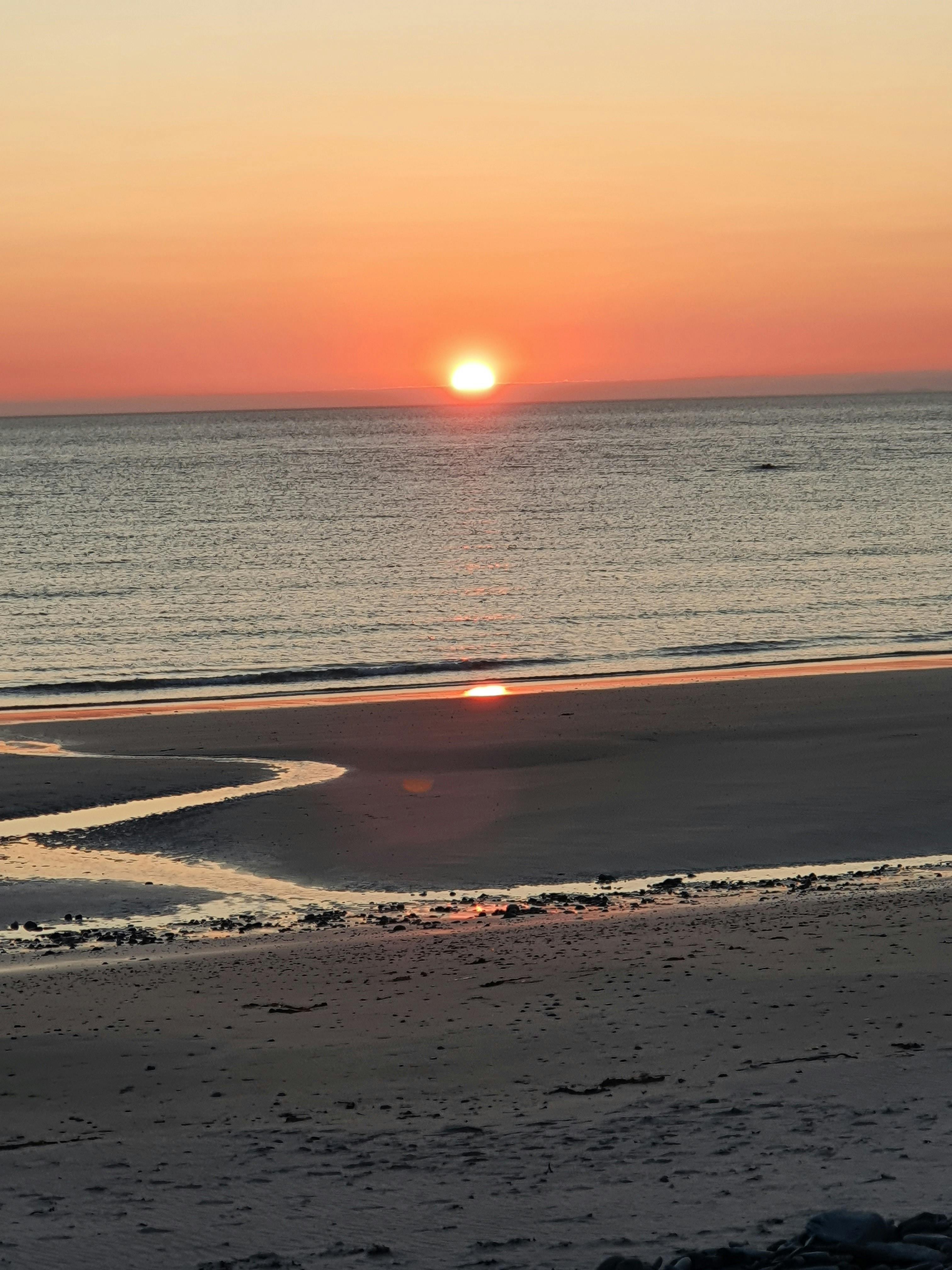Gratis Fredelig strandsolnedgang med livlige oransje fargetoner over havet, som reflekteres på sandkysten. Arkivbilde