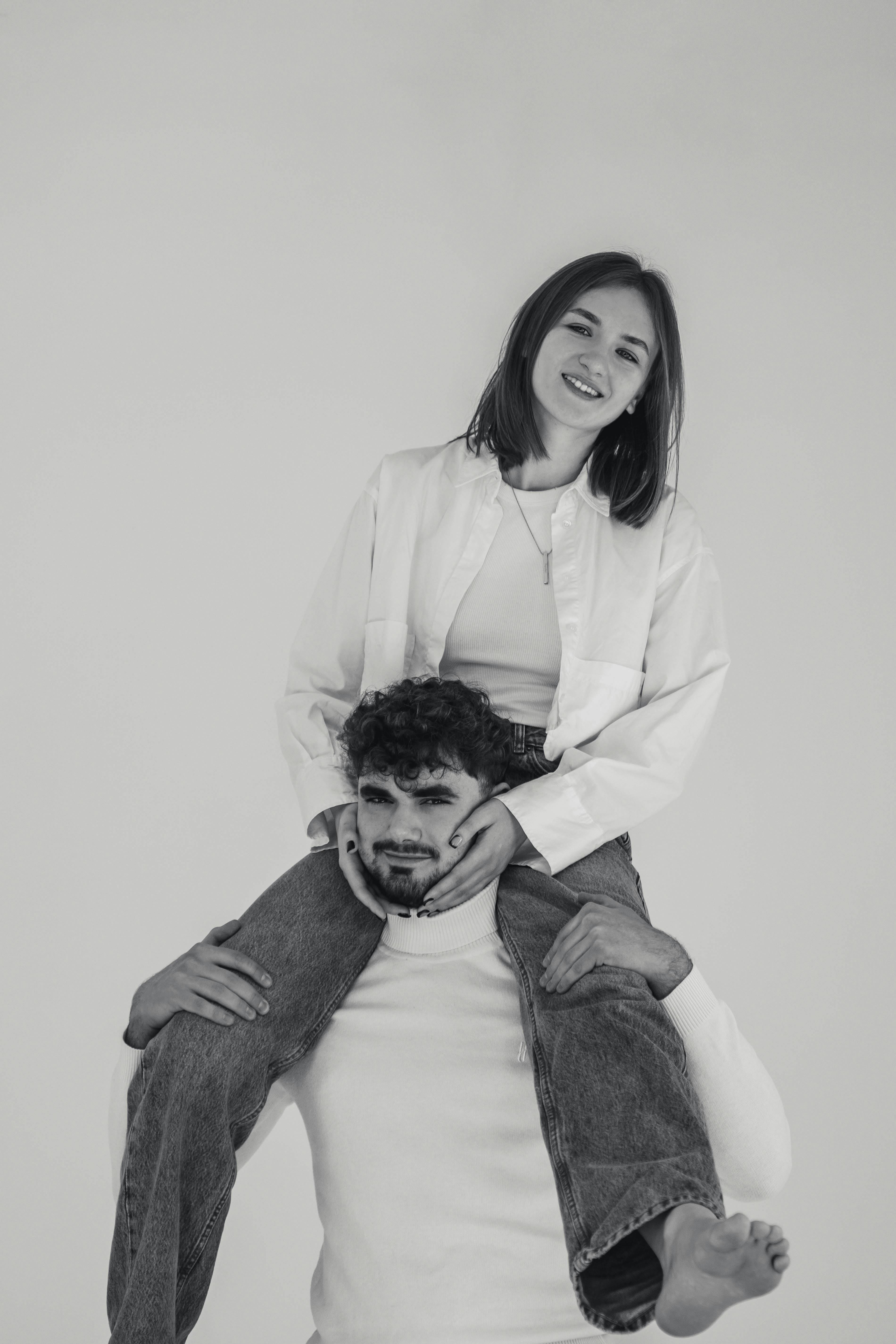 Free A young woman sits on a man's shoulders, smiling in a playful black and white studio portrait. Stock Photo