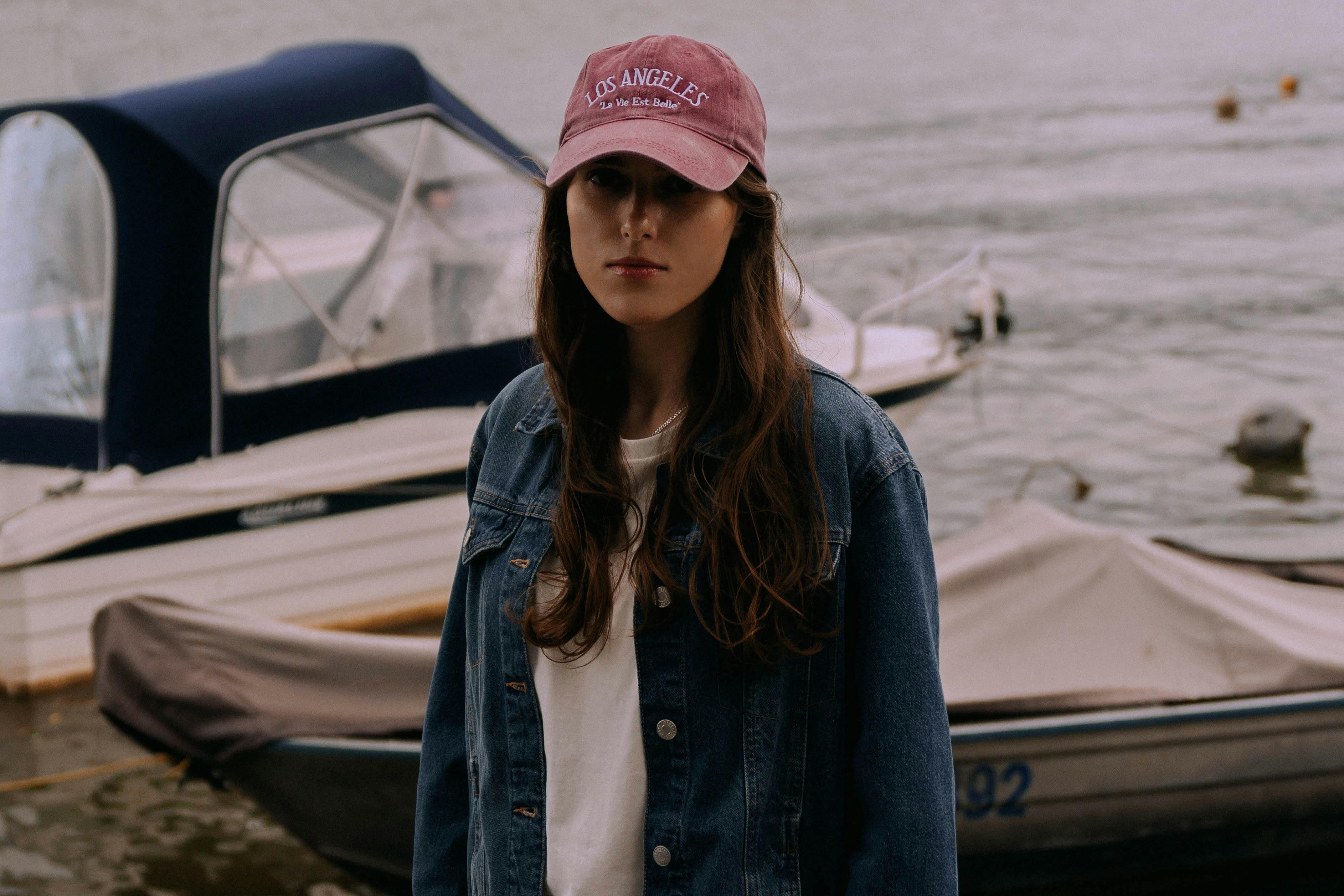 Gratis Seorang wanita muda mengenakan jaket denim dan topi merah muda berdiri di dekat perahu di dermaga pada hari yang mendung. Foto Stok