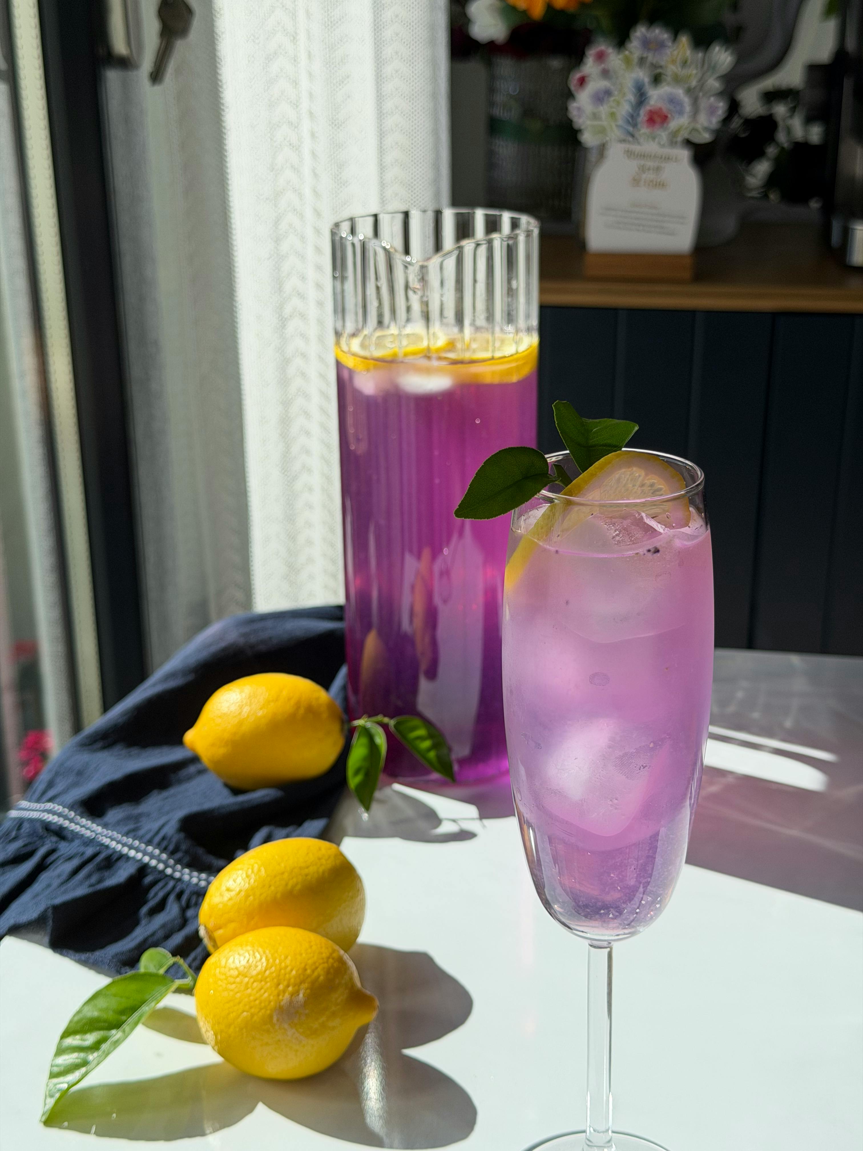 Gratis Segelas dan teko limun lavender dengan irisan lemon segar di atas meja yang terkena sinar matahari. Foto Stok