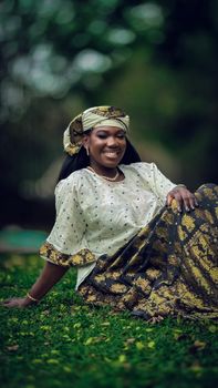 A smiling woman in beautiful traditional clothing sits on green grass outdoors.