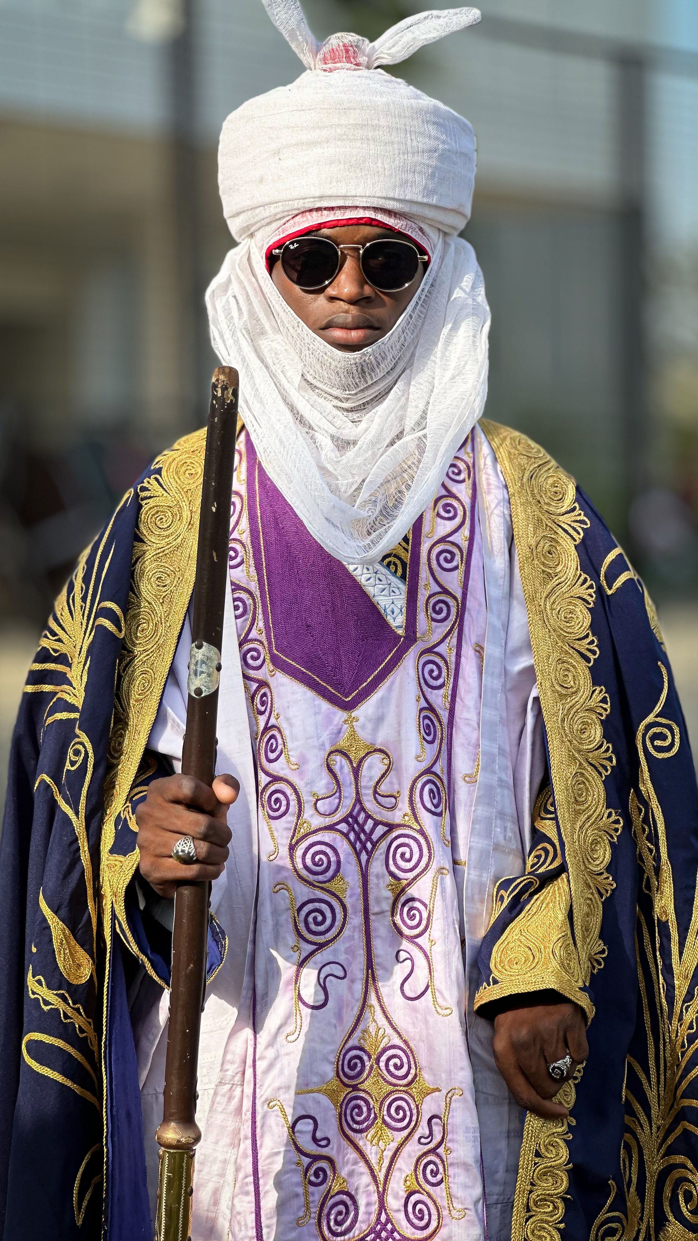 A regal African figure wearing traditional attire and holding a staff, captured outdoors in bright daylight.