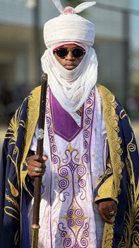 A regal African figure wearing traditional attire and holding a staff, captured outdoors in bright daylight.