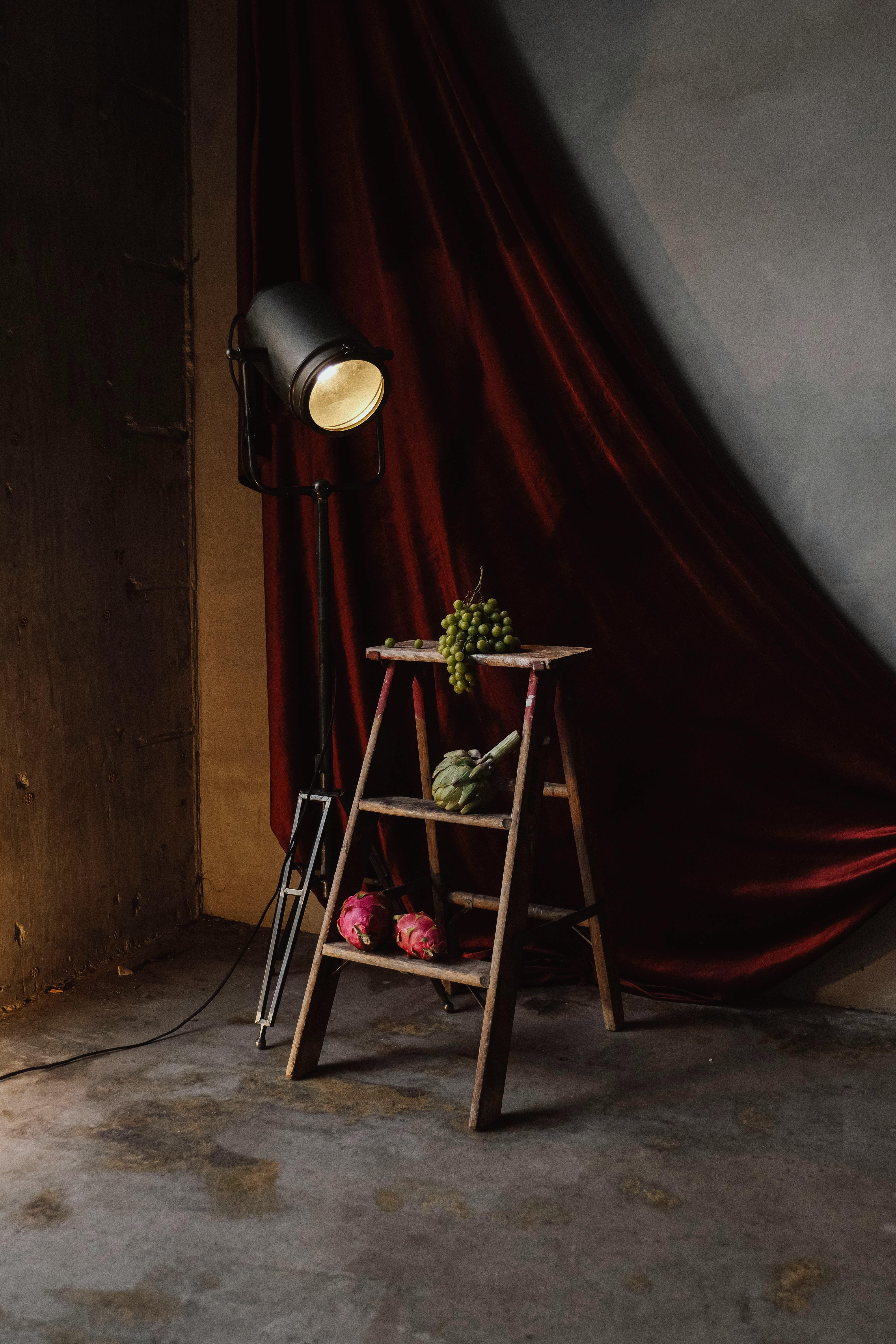 Kostnadsfria Vintage-uppsättning med druvor på en trästege, bredvid en gammaldags spotlight mot en röd draperad gardin. Stock foto