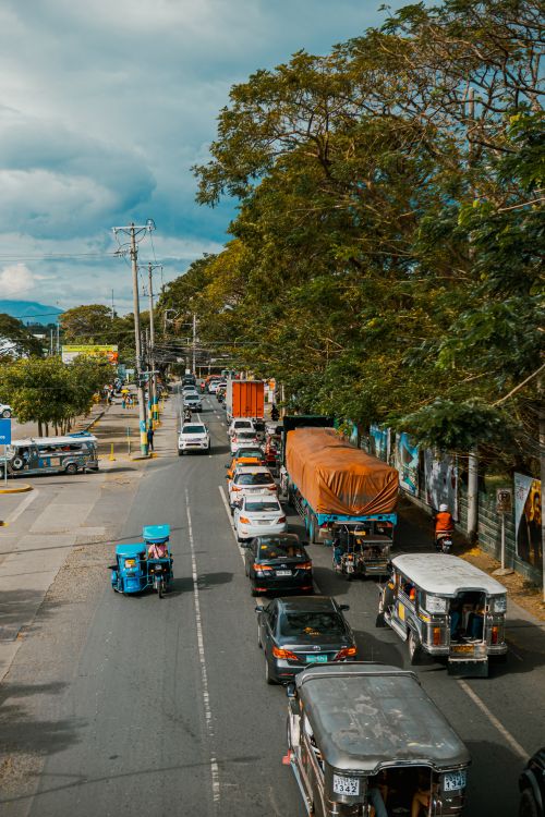 Free A bustling urban street scene with tricycles and cars under a bright sky. Stock Photo