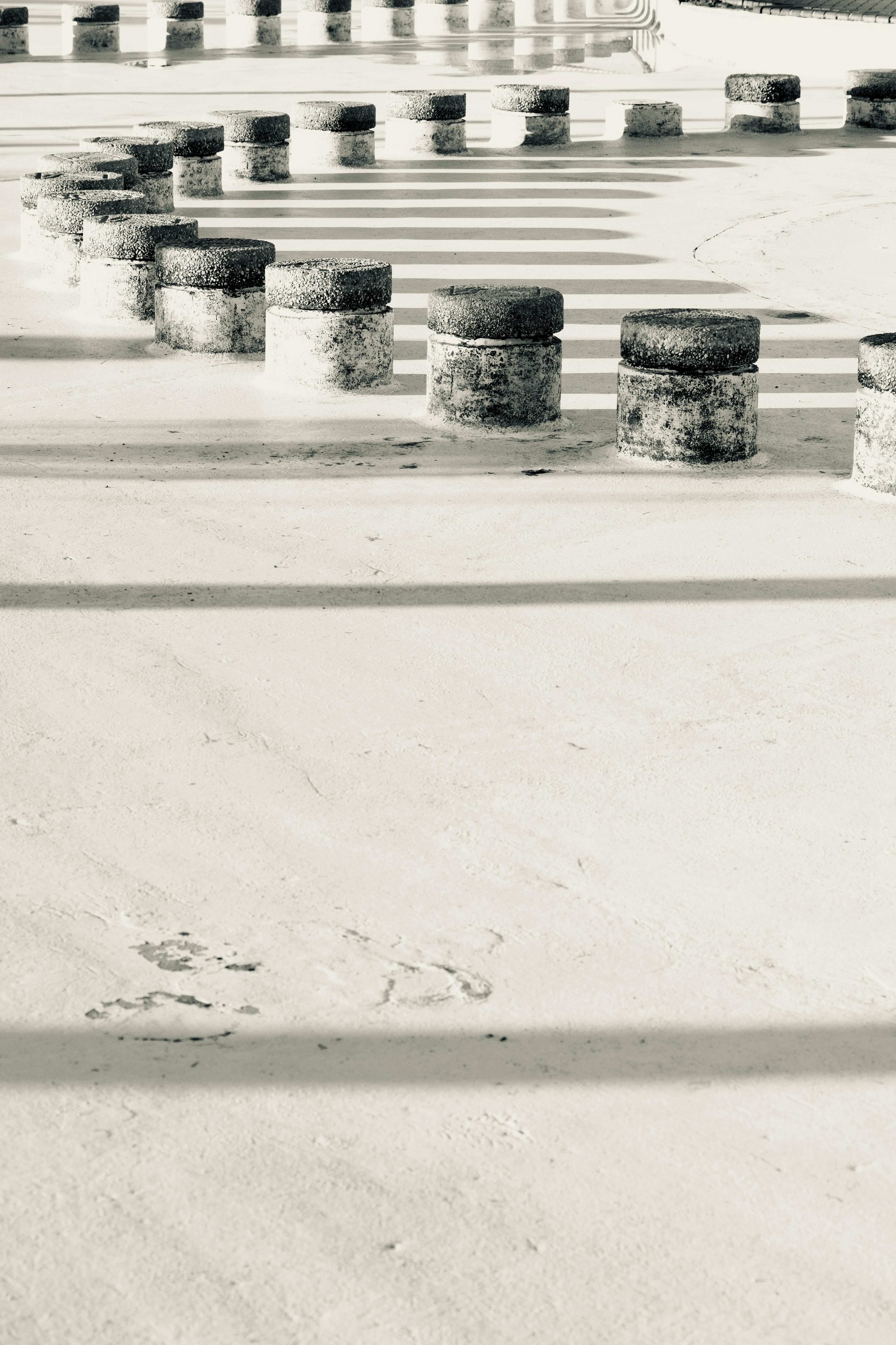 Gratis Immagine monocromatica di cilindri geometrici in cemento che proiettano lunghe ombre su una superficie urbana. Foto a disposizione