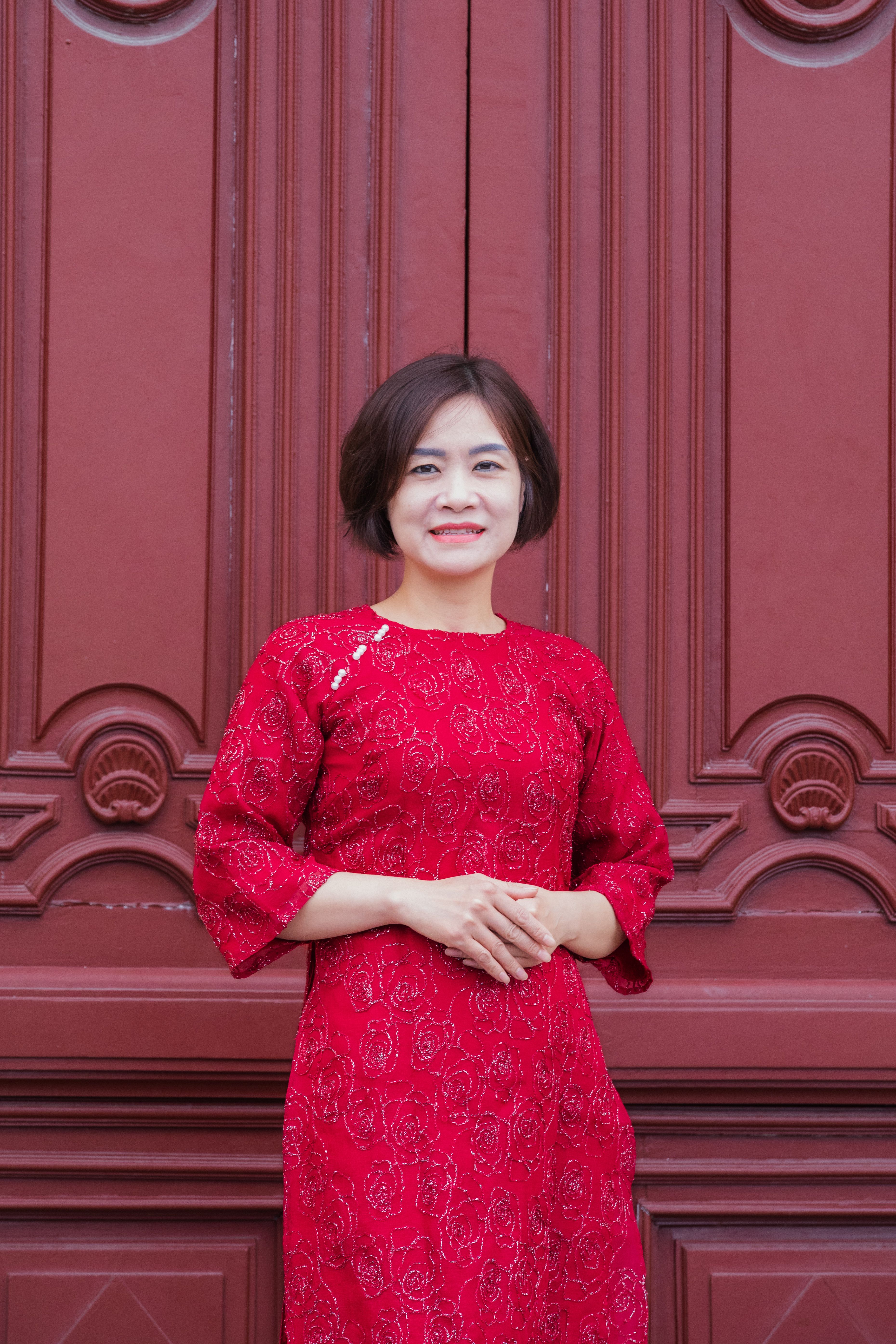 A woman stands confidently in front of a decorative door, wearing a traditional red dress.