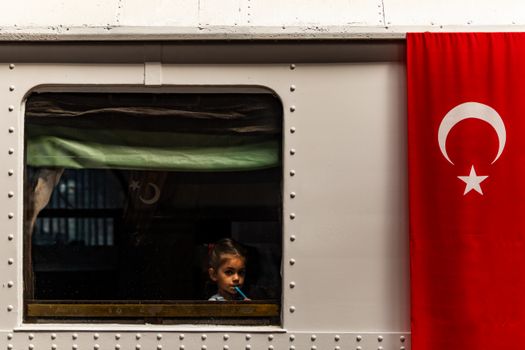 A young girl looks out from a train window adorned with a Turkish flag, evoking a travel narrative.