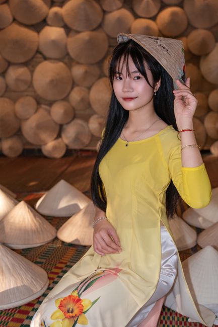 Woman in yellow áo dài posing with a conical hat in a cultural setting.