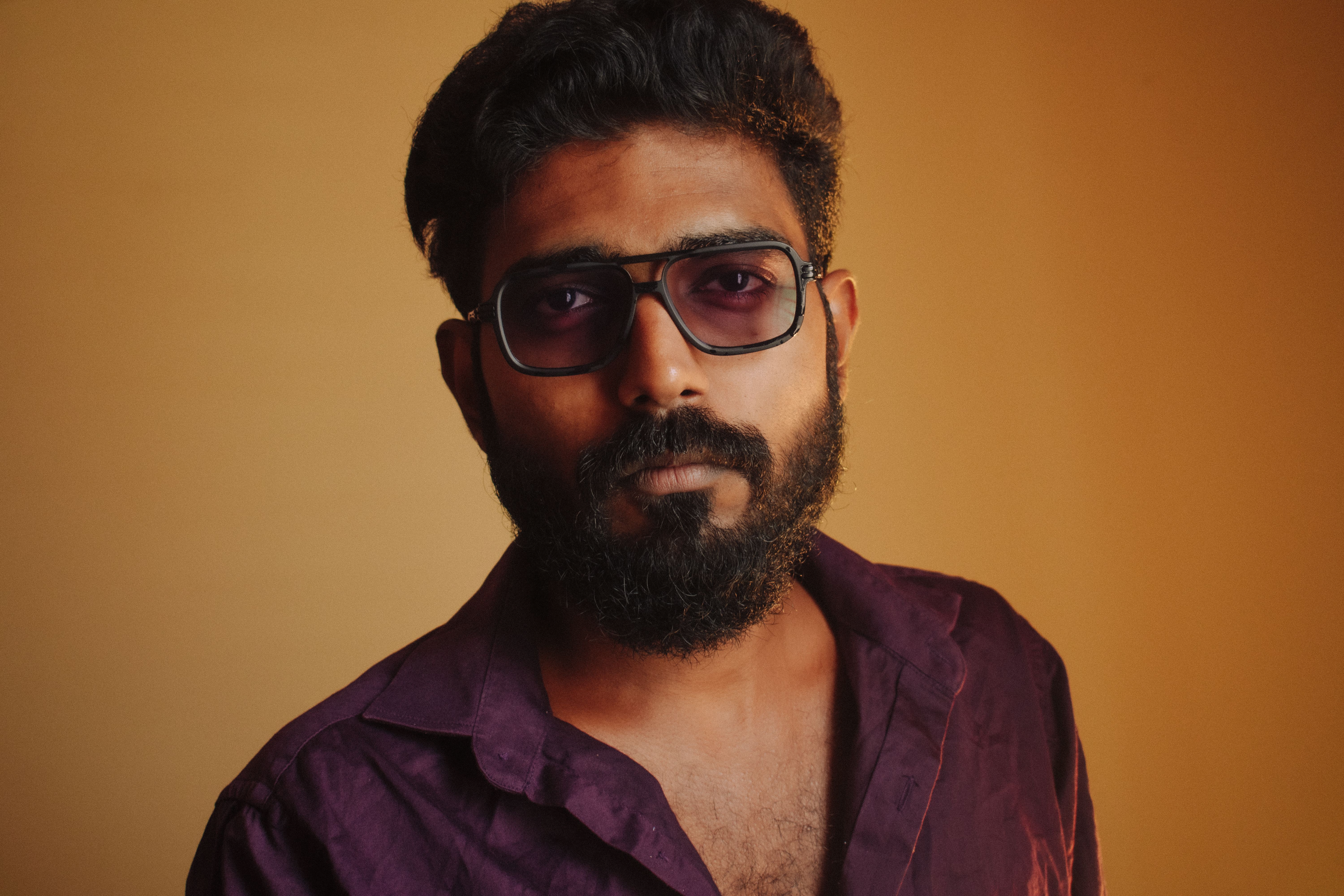 A close-up portrait of an adult man with a beard wearing sunglasses and a purple shirt.