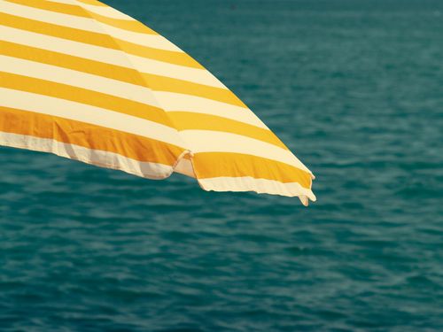 Free Yellow and white striped beach umbrella against blue ocean backdrop, evoking summer vibes. Stock Photo